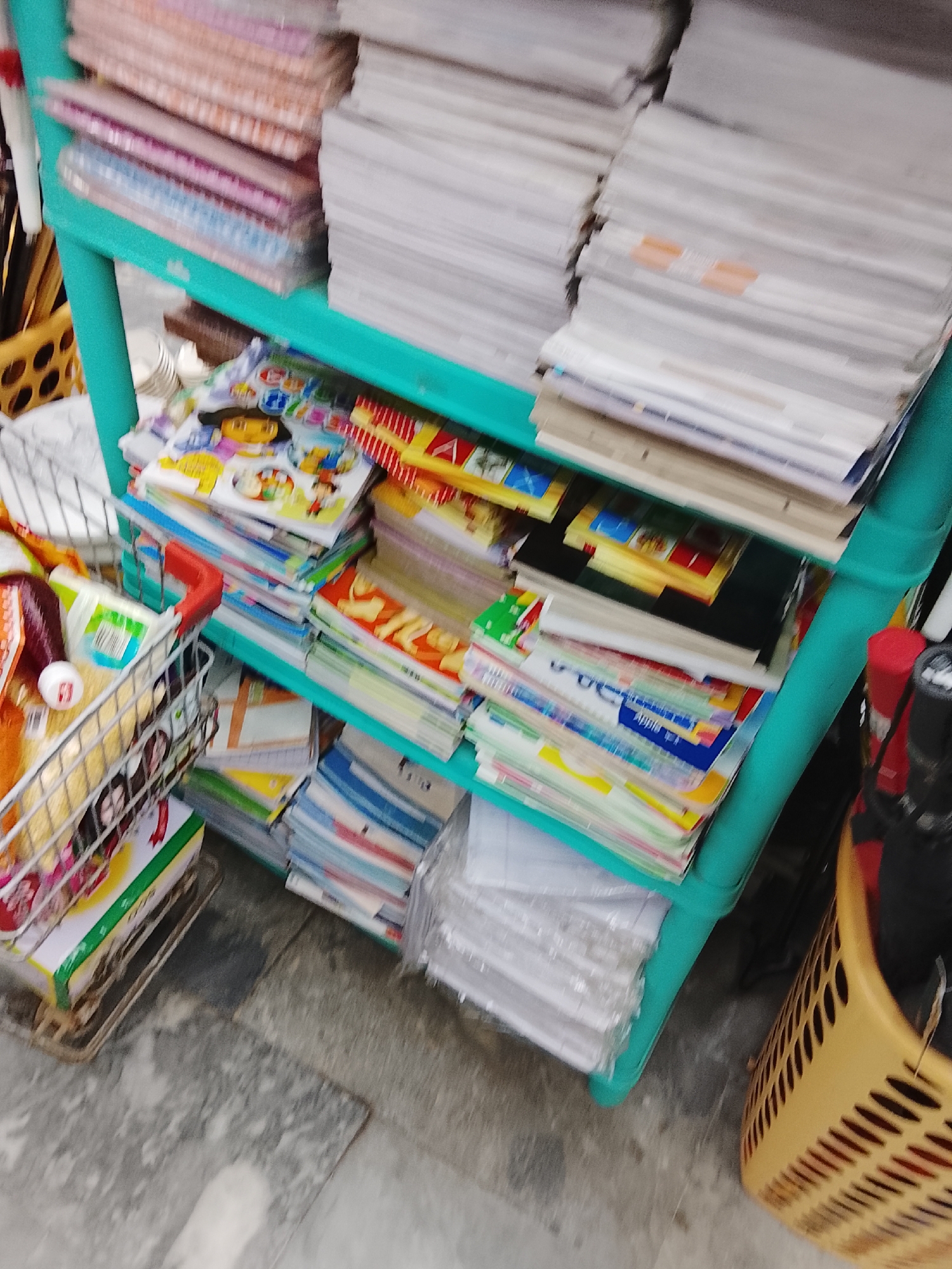 A blurry, medium-angle shot shows a turquoise shelving unit in what appears to be a shop in Gulgasht Colony, Pakistan. The unit is filled with stacks of paper, notebooks, and children's books. The top shelf contains large stacks of white paper and some notebooks with pink and blue covers. The second shelf has stacks of children's books with colorful covers, featuring cartoon characters. The third shelf holds more notebooks and stacks of paper. To the left of the shelving unit, there is a wire shopping basket filled with groceries and a green box. To the right, there is a tan basket with black objects inside. The floor is gray. The overall impression is of a cluttered retail space.