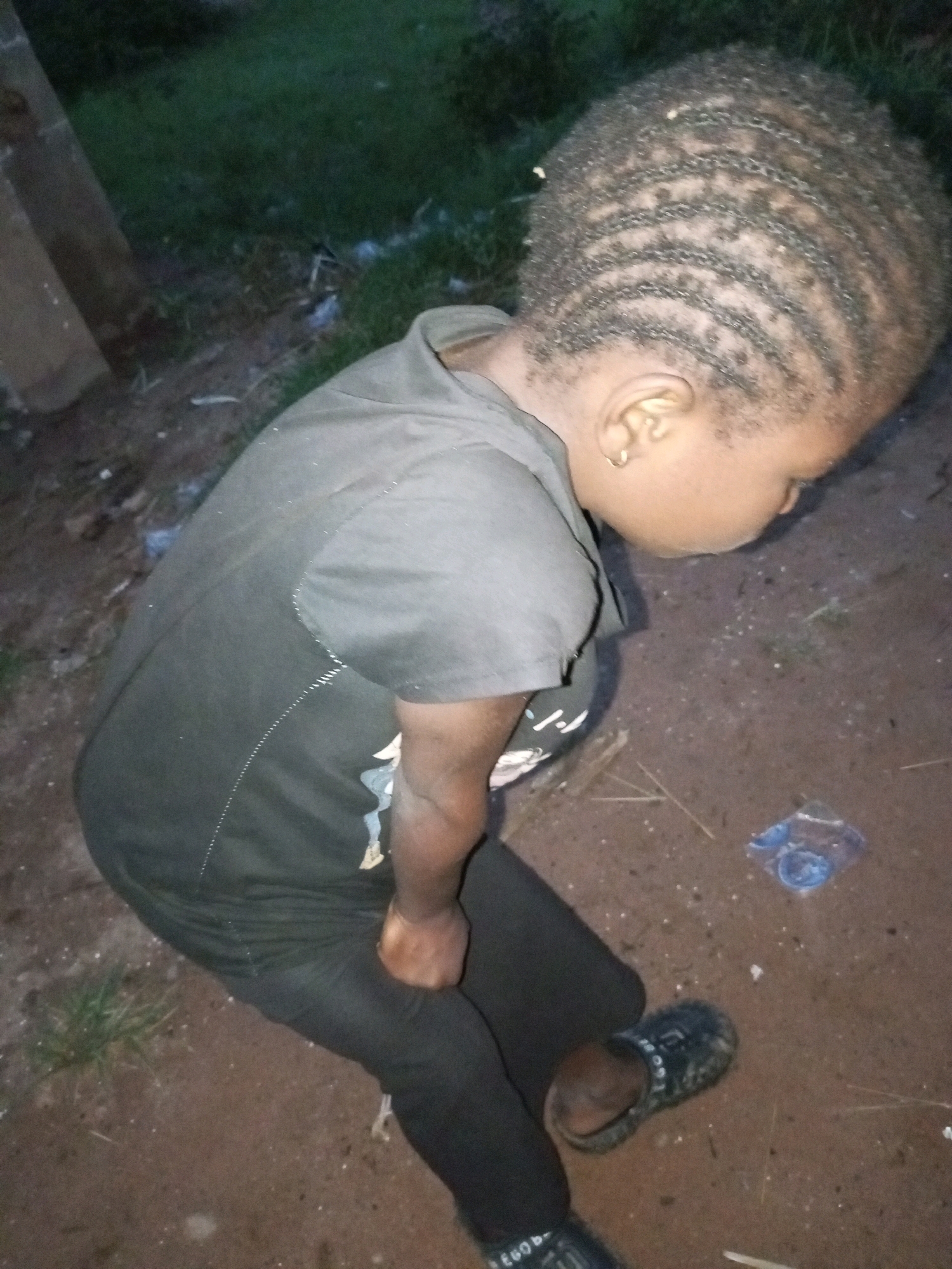 In a dimly lit outdoor scene, a young child with intricately braided hair is seated on the ground. The child is wearing a dark t-shirt and dark pants, with dark-colored sandals on their feet. The ground appears to be a mix of dirt and sparse grass. In the background, a wooden object is visible, along with more grass. A small blue object, possibly a wrapper or a toy, is on the ground near the child. The overall lighting suggests it might be evening or nighttime. The location is identified as Tempure, Nigeria.