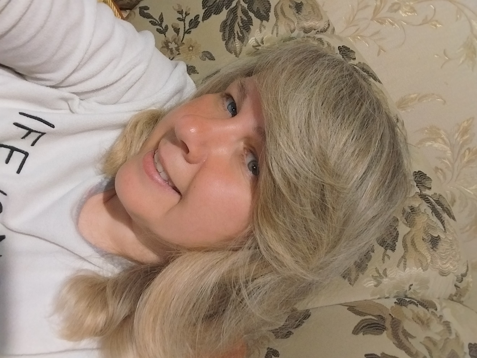 A woman with blonde hair and blue eyes is captured in a close-up, upward-angled shot, smiling directly at the camera. Her head is tilted, suggesting she is lying down, likely on a couch or bed. She has fair skin and a relaxed, happy expression, with her teeth slightly visible as she smiles. She is wearing a white, textured long-sleeved top, and the word "LIFE" is partially visible in black lettering on her chest. The background consists of a patterned fabric or wallpaper with an intricate floral and foliate design in shades of beige, cream, and darker brown, contributing to a cozy indoor ambiance. The lighting is soft and even, characteristic of an interior setting. The scene conveys a moment of ease and contentment, possibly within a home in Orenburg, Russia.