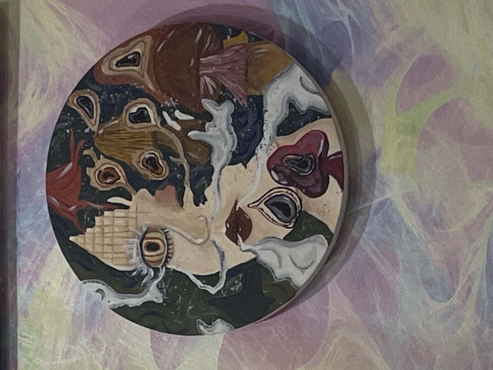 A circular, painted artwork is affixed to a wall. The artwork depicts a surreal, abstract face with elements of mushrooms and smoke integrated into its design. The face is predominantly rendered in light skin tones, with a prominent, stylized eye. Dark red lips are also visible. Surrounding the face are various mushroom shapes in shades of brown, red, and yellow, some appearing to sprout from or merge with the facial features. Wisps of white smoke or vapor curl around the face, adding to the ethereal and dreamlike quality of the piece. The background of the artwork is a dark, textured blue or black, with specks of what might be stars or abstract patterns. The wall behind the artwork has a soft, pastel-colored, textured pattern, appearing to be a gradient of pale pinks, yellows, and blues with a subtle mottled or watercolor-like effect. The artwork is centrally positioned on the wall, and no other objects or people are visible in the frame. The location is identified as ЖК «Борисоглебское», Russia.