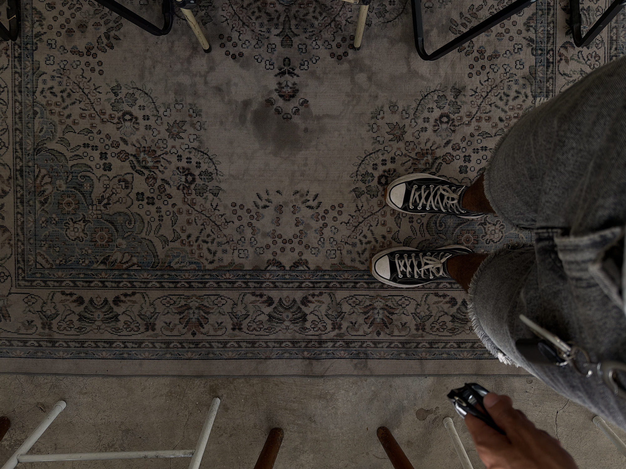 The image presents an overhead view of a person standing on a patterned rug, presumably indoors. The person is wearing black Converse sneakers and light-wash denim overalls. They hold a black car key and a key chain in their right hand. Keys dangle from the right pocket of the overalls. Parts of furniture, likely chairs, are scattered around the frame's edges. The rug has an intricate design with floral and geometric patterns in faded blues, browns, and grays. Below the rug, a concrete floor is visible, partially covered by furniture legs. There is no definitive evidence in the image to confirm it was taken in North Kuta, Indonesia.
