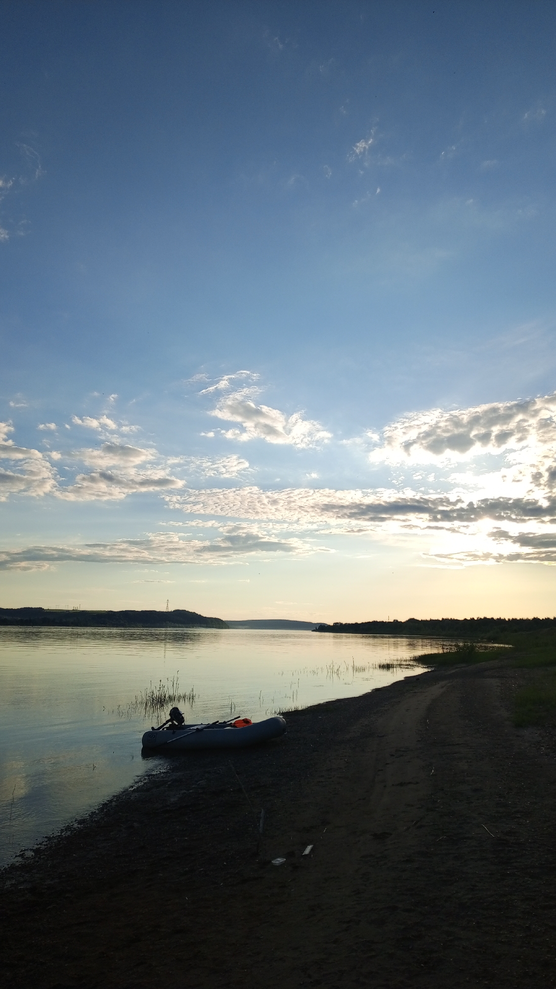 The image depicts a tranquil, uninhabited riverside landscape in or near Шолья, Russia, observed during either dawn or dusk. The scene is bisected horizontally by a wide, calm river, with a prominent sky above and a dark, sandy bank in the foreground.
The upper portion of the frame features a vast sky, transitioning from a light blue at the zenith to a pale, golden-blue hue near the horizon. Scattered stratocumulus and altocumulus clouds are present, with a dense cluster on the right partially obscuring the low-angle sun, which emanates a strong, bright glare.
The wide river displays a smooth, reflective surface, mirroring the sky and the sun's golden light. Across the water, a distant landmass forms the opposing bank, covered with dense, dark green foliage, indicative of a forested area. On the far left of this distant bank, two slender, dark vertical structures, likely power pylons, are visible against the horizon.
The immediate foreground features a dark, damp sandy or muddy bank that slopes gently into the river. This bank appears to be an unpaved track, displaying faint impressions consistent with tire or foot traffic. Sparse, thin reeds emerge from the shallow water near this bank.
A grey inflatable boat, equipped with an outboard motor, is positioned partially in the water and partially on the sand in the lower-left quadrant. Oars are stowed within the vessel, alongside several orange objects, identified as life vests or similar flotation devices. A singular, thin wooden stick is embedded vertically in the sand adjacent to the boat. No human figures are present in the depicted environment.
