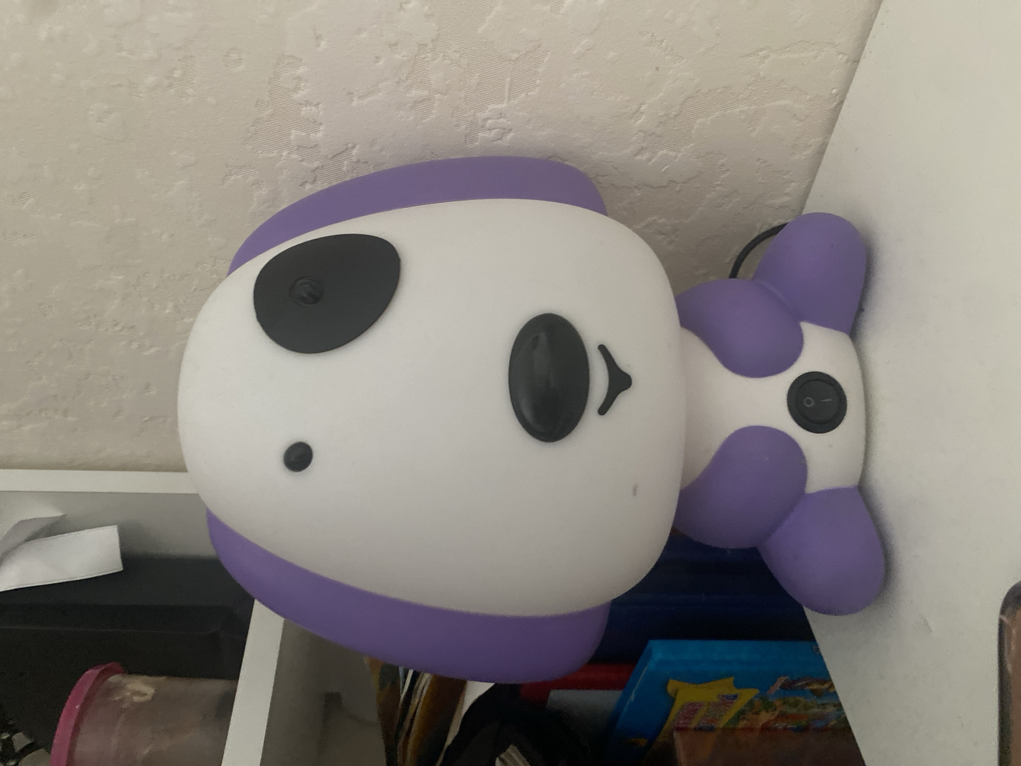 An interior shot shows a white and purple dog-shaped night light on a white surface. The night light has black oval spots for eyes, a black oval nose, and a black curved line for a mouth. A black power switch is located on its chest. The night light is positioned in front of a textured, cream-colored wall. To the left of the night light, a white shelf is visible, holding a pink-lidded container and some books. The scene does not provide any information about the city of Novosibirsk, Russia.