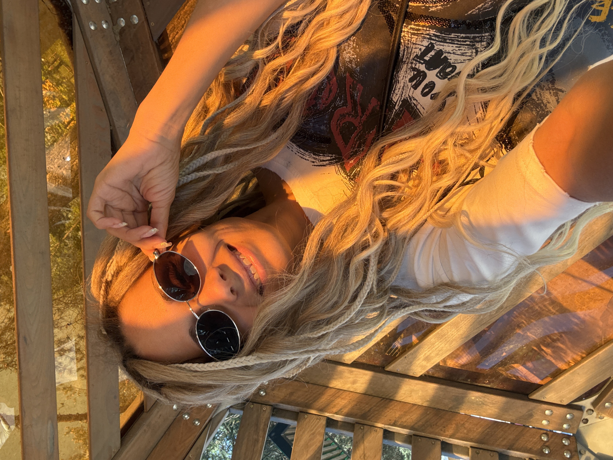 A woman with long blonde hair, styled with braids, is seen wearing round sunglasses and a light-colored top under a black graphic vest. She is smiling and holding the sunglasses frame with her left hand. The surrounding environment appears to be a wooden structure, potentially a lookout or enclosed area, with glimpses of greenery through openings, potentially indicating a park or natural setting. While the location is specified as Шебунино, Russia, no specific geographical or urban indicators are present to confirm that location within the image.