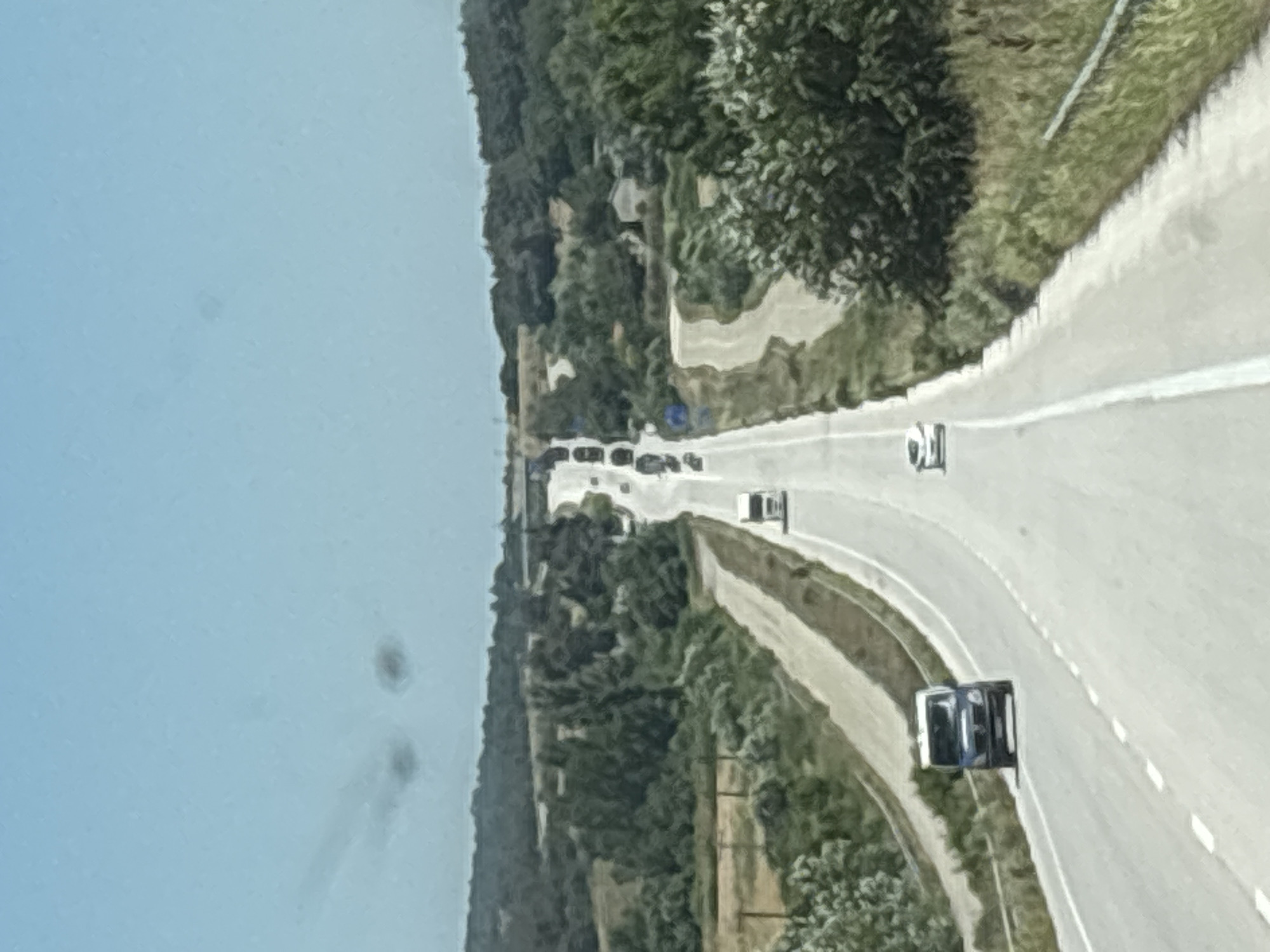 The image depicts a section of a highway in Λύρα, Greece, under a clear, pale blue sky. The road curves and ascends into the distance, flanked by lush green trees and vegetation on either side. Several vehicles are visible on the highway. In the foreground, a blue van is traveling away from the camera. Further up the road, a white car is also heading away. A white truck is seen further ahead, and a procession of other vehicles, including trucks and cars, stretches along the road in the distance. The landscape suggests a rural or semi-rural environment. The quality of the image is slightly blurred, possibly due to motion or lighting conditions.