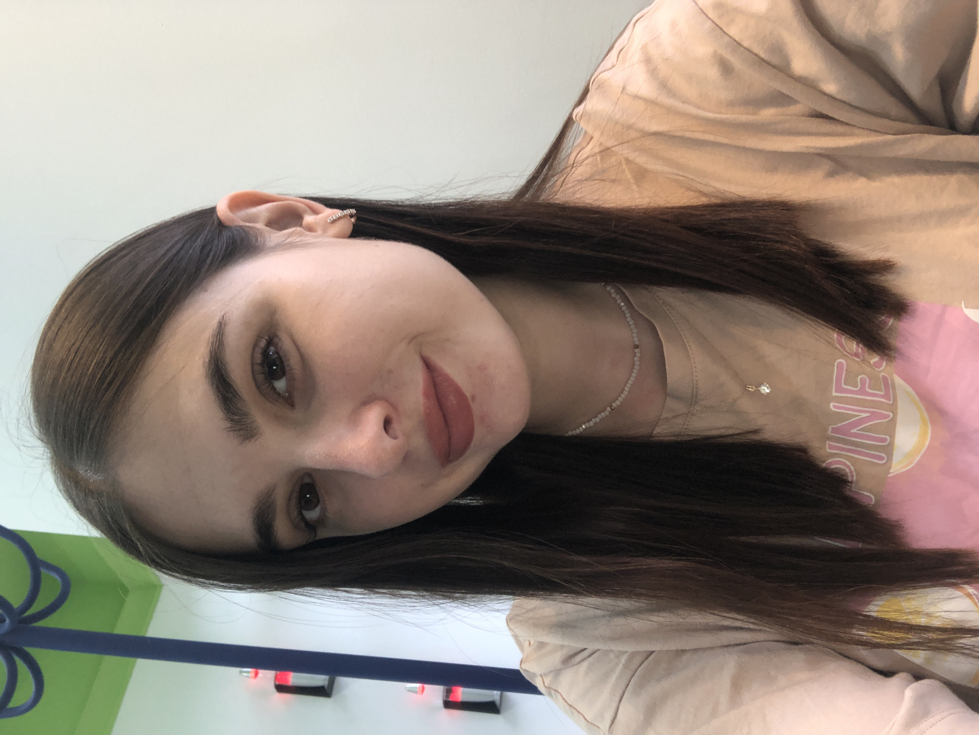 A young woman with long, dark brown hair and fair skin is posing for a selfie. She is wearing a light peach-colored t-shirt with a graphic design that includes the word "PINES" in pink lettering. She has on a delicate necklace with a small pendant and a hoop earring in her left ear. Her makeup includes lipstick in a rosy-brown shade. The background appears to be an indoor setting with a light-colored wall and a green ceiling with a blue decorative element. The image is cropped to focus on her face and upper torso. There is no indication of the city of Chaikovsky, Russia, within the visual information provided.