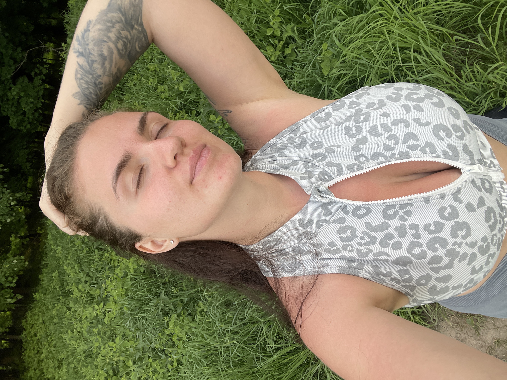 A woman lies on her back in a grassy area. She has brown hair, and fair skin. She is wearing a gray and white patterned sports bra with a zipper in the front. Her eyes are closed. One arm is bent at the elbow and positioned above her head. Her left arm has a large tattoo. The green grass surrounds her. The scene appears to be outdoors. The location of the photo is not discernible as Ostrov, Russia from this image.