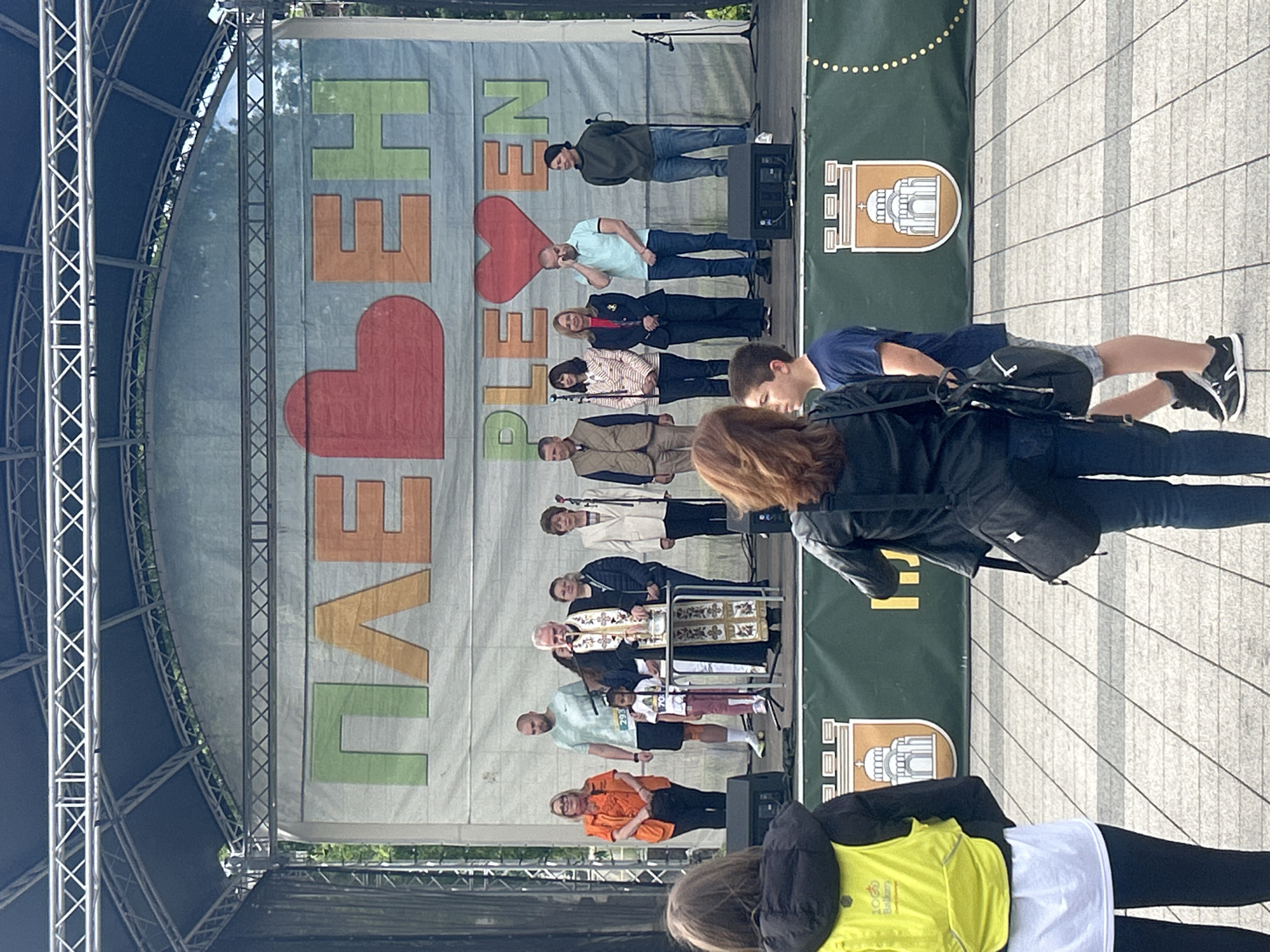 The image shows a group of people on a stage in front of a banner that reads "LIVE <3 PLEVEN." The stage is located in the city of Pleven, Bulgaria, indicated by the coat of arms on the banner. There are at least 10 people standing on the stage, including a priest in a traditional robe, with some people positioned behind podiums and microphones. The stage is constructed with metal framework, and there is a paved area in front of the stage with people standing in the foreground. The banner uses a color scheme of green, orange, and red. The background is obscured by the banner and the stage framework, but some foliage is visible. The photograph appears to be taken from the audience's perspective.
