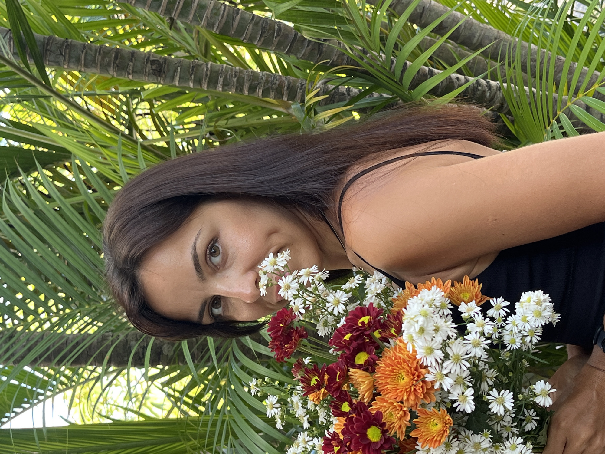 This portrait-oriented image, rotated 90 degrees counter-clockwise, features a woman holding a vibrant bouquet of flowers against a tropical backdrop. The woman, with long, dark brown hair and brown eyes, looks directly at the camera with a gentle, soft smile. She wears a black top, with a thin strap visible on her shoulder. Her head is slightly tilted, and some small white flowers from the bouquet are positioned near her nose, suggesting she is smelling them or playfully posing.
The bouquet is rich in color, primarily featuring white daisy-like flowers, deep red/maroon blooms, and orange chrysanthemum-like flowers. Her left hand, with a partially visible dark-colored watch on the wrist, holds the base of the bouquet.
The background is filled with the lush green fronds and distinctive textured trunks of palm trees, strongly indicating a tropical or garden environment. The bright, natural lighting filtering through the leaves suggests it is daytime, under sunny conditions. The woman's calm and pleasant expression contributes to a serene mood. There is no visible text. The location context is Kuta Selatan, Indonesia.
