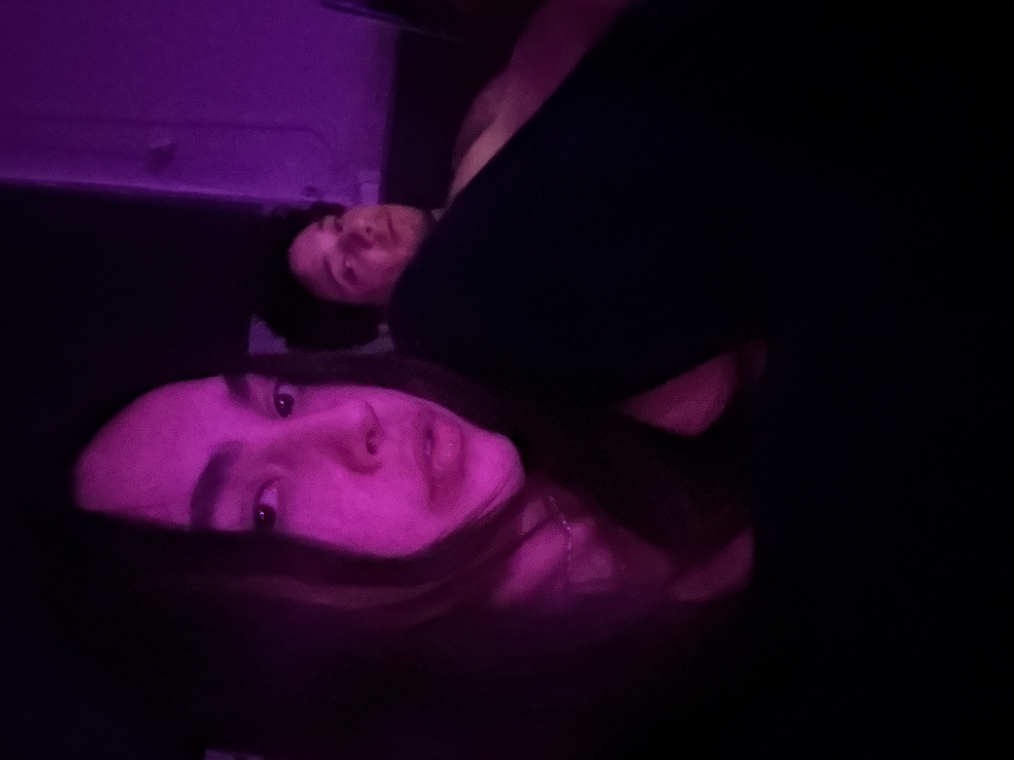 Two individuals are visible in the image. One is a woman, centered in the foreground, with fair skin and long, dark hair. The other is a man, positioned behind and slightly above the woman, also with fair skin and short, dark hair. The lighting is primarily dark with a purple hue, which affects the color of the skin and surroundings. A dark object, possibly a piece of clothing, is prominently placed in the middle of the image, partially obscuring the man. The background appears to be an interior space with a light-colored wall. The location cannot be determined from the image.