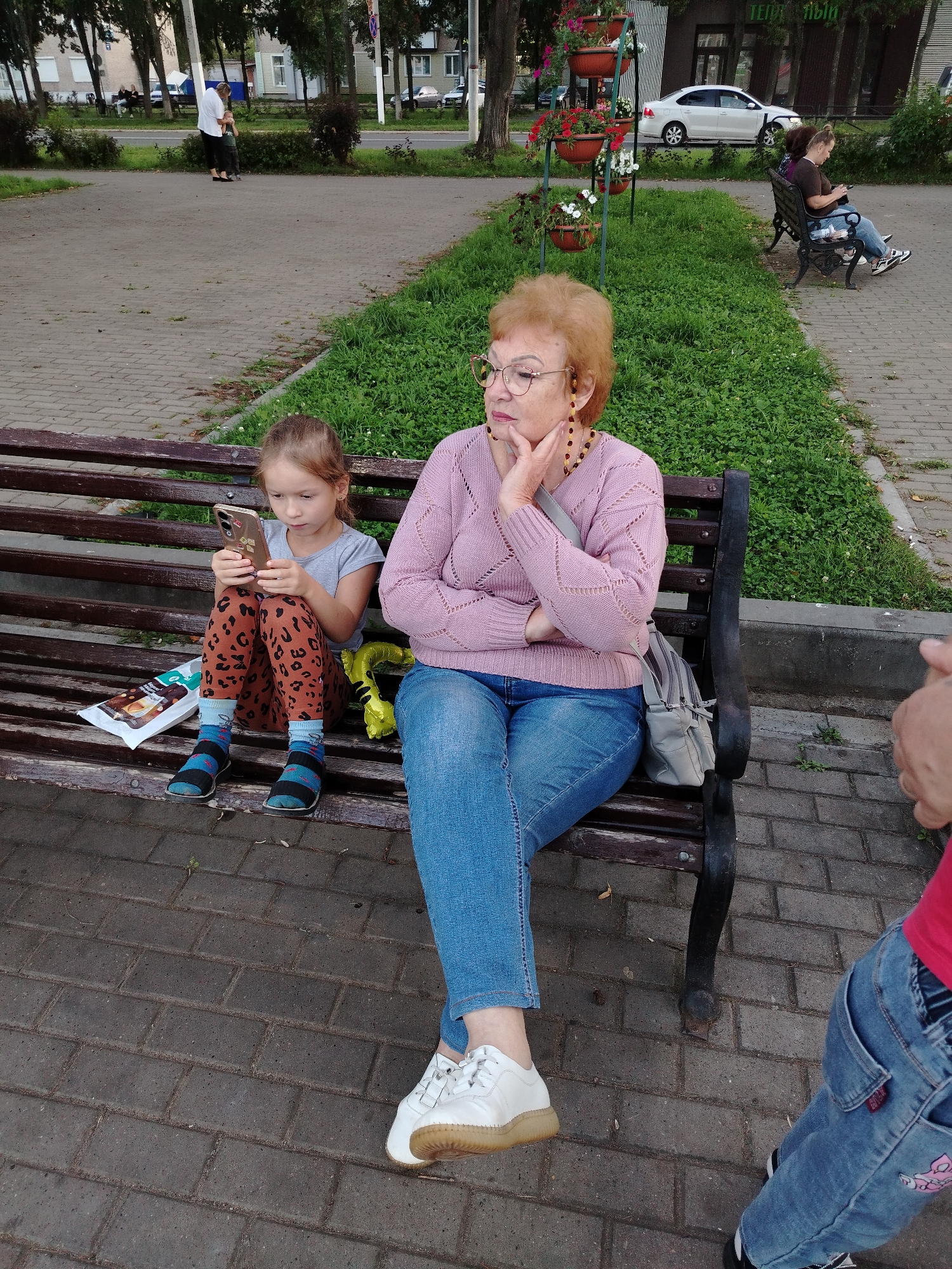 In the city of Chaikovsky, Russia, on a brick-paved walkway in what appears to be a park or public square, two individuals are seated on a wooden park bench.
The individual on the left is a young girl, seated with her legs crossed. She is wearing leopard-print leggings, blue and black striped socks, and dark shoes. She is engrossed in using a smartphone, holding it with both hands. Beside her on the bench lies a wrapped snack and a yellow, deflated balloon.
To the right of the girl sits an older woman, wearing glasses, a pink knitted sweater, and blue jeans. She has her hand resting on her chin, appearing contemplative. A light-colored shoulder bag is placed on the bench next to her.
In the background, a grassy area is visible, bordered by trees and a road. On the far side of the road, buildings and parked cars can be seen, including a white sedan. Another woman is seated on a park bench further in the background, also looking at her phone. The overall setting suggests a public outdoor space in an urban environment.
