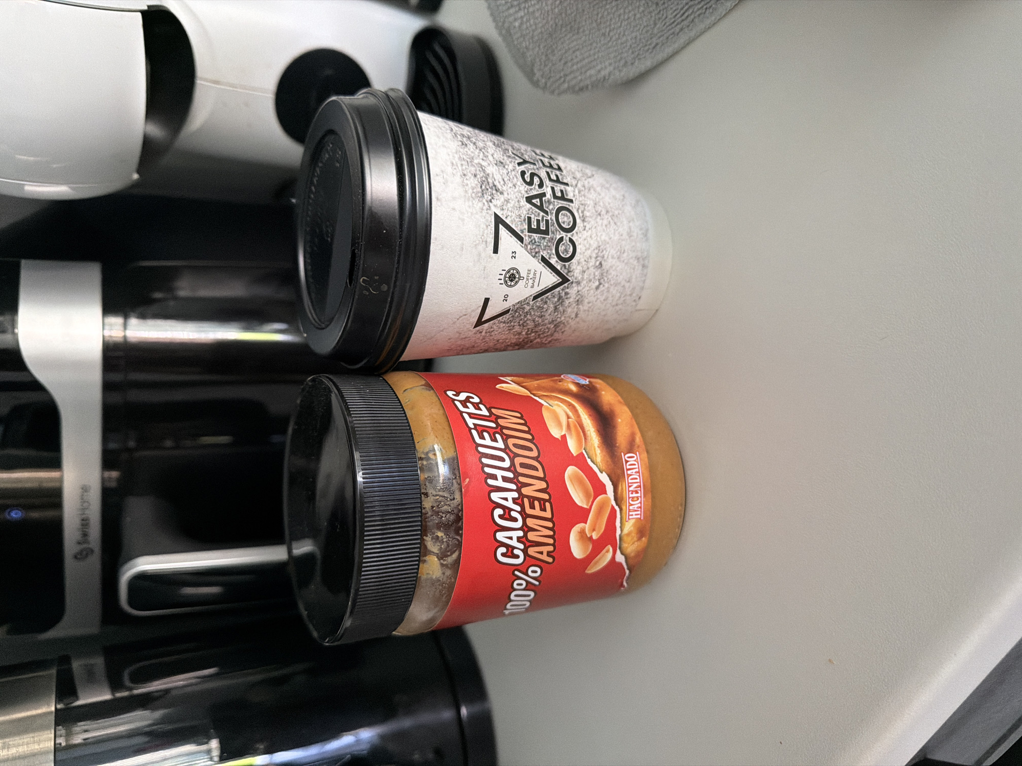 The image shows a close-up, tilted view of a table or surface, with two food items prominently displayed. On the top, a disposable coffee cup with a black lid has a white and grey speckled design with bold black lettering that reads "EASY COFFEE" with a stylized "V" shape. Below the coffee cup is a jar of peanut butter. The jar has a black lid and a red label with white text that states "100% CACAHUETES AMENDOIM" and "HACENDADO" in smaller white lettering. The label also features an image of peanuts. The background shows a white appliance, possibly a coffee maker or similar kitchen appliance, and a textured grey fabric. The lighting suggests an indoor setting, likely during the day. The overall impression is of everyday items on a surface, possibly in a kitchen or break room environment. The location context provided is Finestrat, Spain, which might indicate where these items are commonly found or purchased. There are no people or animals visible in the image, and no specific activity or event is depicted other than the arrangement of the objects. The focus is on the textures and branding of the coffee cup and peanut butter jar.