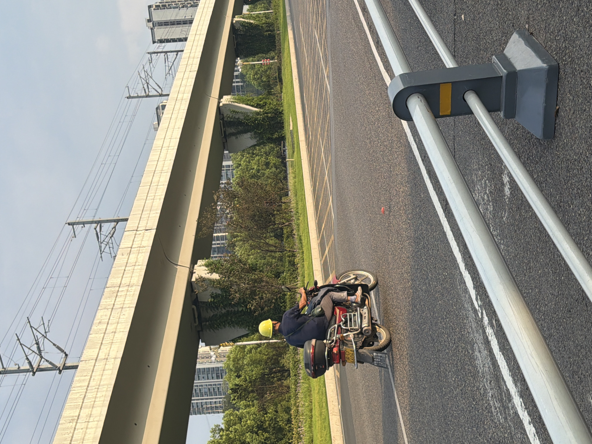 A man wearing a yellow hard hat and a dark blue shirt rides a red motorcycle on a paved road in Jiangjin, China. The motorcycle has a black storage box on the back and a license plate with the characters "66064". The road is lined with white and yellow markings. In the background, a concrete elevated structure, possibly a bridge or overpass, runs parallel to the road. This structure has overhead power lines and is supported by concrete pillars covered in ivy. Beyond the elevated structure, there are trees and tall buildings, indicating an urban or suburban setting. The sky is clear and blue with some wispy clouds. A silver guardrail with dark gray supports is visible in the foreground, partially obscuring the road. The lighting suggests it is daytime, likely late afternoon due to the angle of the sunlight.