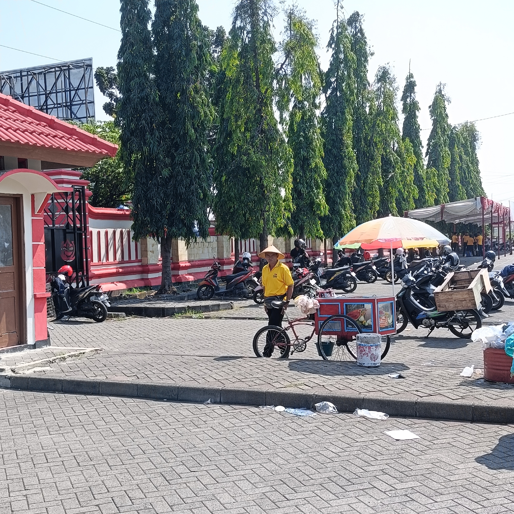 The image captures an outdoor scene in the city of Kendal, Indonesia, likely in a public space such as a market or parking area. In the foreground, a street vendor wearing a conical hat and a yellow shirt stands next to a red and white pushcart, possibly selling food or drinks. The cart has a colorful display and appears to be set up for business. Behind the vendor, a line of parked motorcycles is visible, some with riders wearing helmets. The background features tall, slender trees that line a red and white striped wall. A building with a red roof and white trim is visible to the left, and a large billboard structure is seen in the distance. The ground is paved with interlocking bricks, and there is some scattered litter, including plastic bags and wrappers, along the curb. The overall atmosphere suggests a warm, sunny day in a bustling urban environment.