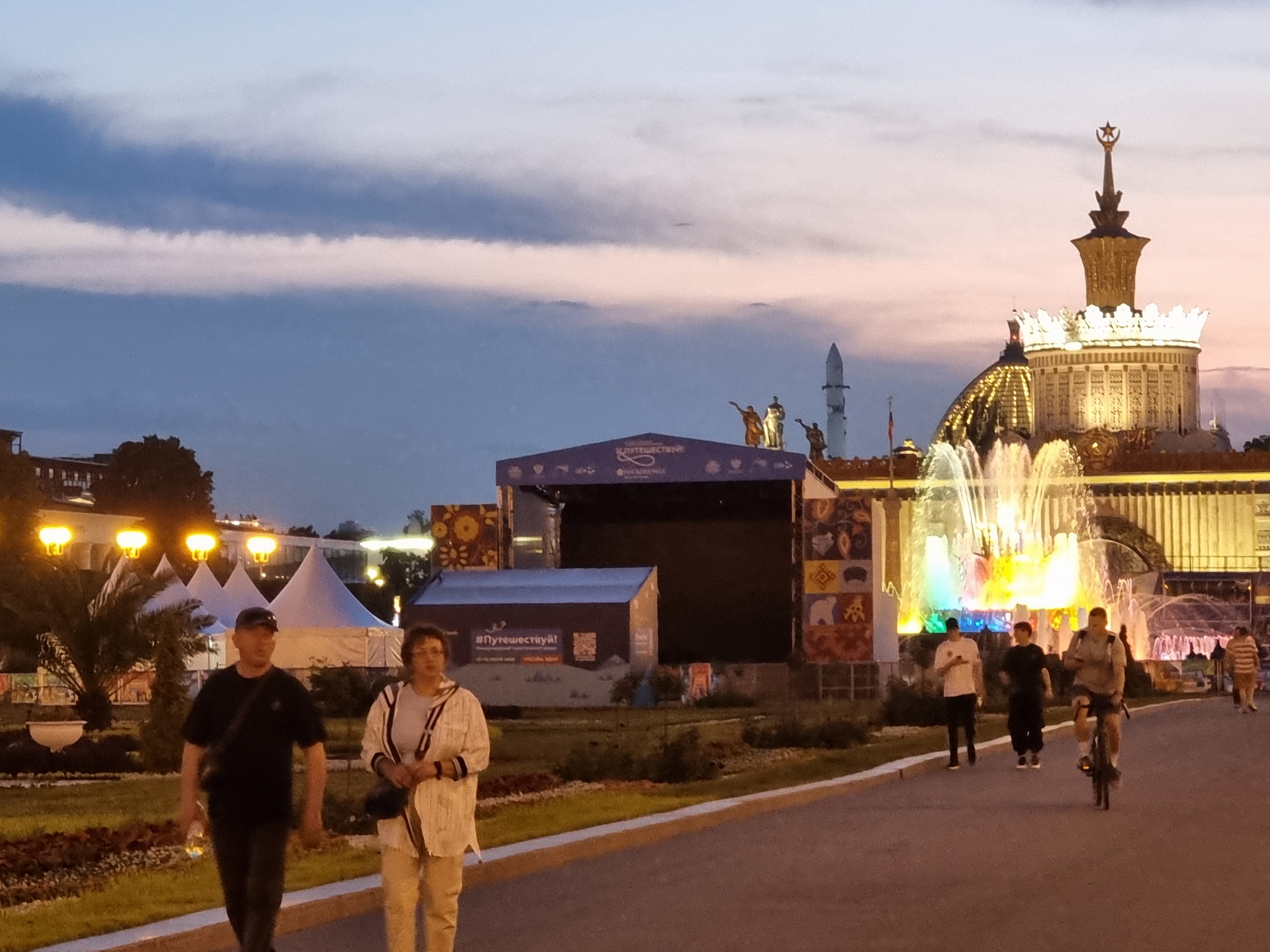 This image captures an evening scene at VDNKh (Exhibition of Achievements of National Economy) in Moscow, Russia. In the foreground, a man wearing a black t-shirt and a cap walks to the left, holding a clear bottle. To his right, a woman in a white jacket with black stripes walks towards the left, holding a black bag. Behind them, a paved pathway runs to the right, where a man on a bicycle is seen cycling to the right. Further behind, several people are walking, and a vibrant, illuminated fountain is active. In the background, a stage structure is set up with a large black screen and colorful decorative panels. Beyond that, the iconic Moscow skyline is visible with illuminated buildings and a towering spire topped with a star. The sky above is a gradient of dusky blues and pinks, indicating the late hour. White tents and palm trees are also present in the middle ground.