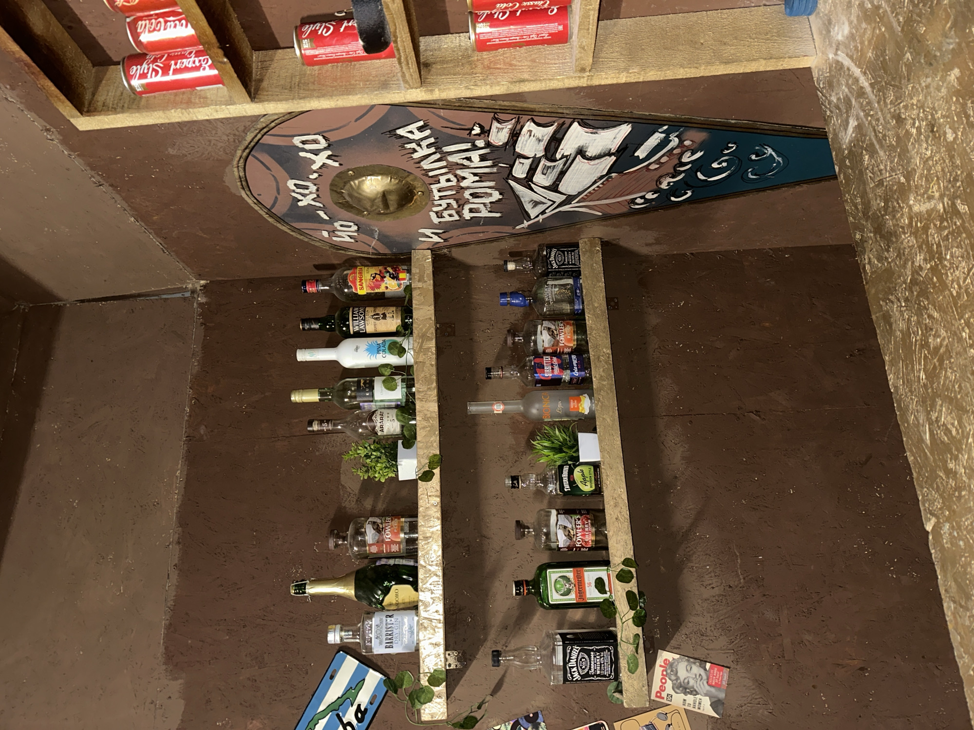 The image displays an indoor bar area situated in Saratov, Russia. The walls are a dark brown, textured finish. Two light-colored wooden shelves are affixed to the left wall. The upper shelf holds a blue and white decorative plate reading "CUBA," a Barrister Dry Gin bottle, a green bottle, a Fowler's Peach spirit bottle, a small artificial potted plant, an Ararat brandy bottle, and two other bottles with partially obscured labels. The lower shelf displays a Jack Daniel's Tennessee Whiskey bottle, a Jagermeister bottle, a Fowler's Cherry spirit bottle, a Tavern Hound Apple Style bottle, a second artificial potted plant, a Bionica liquid bottle, and a SBITRITTEB Cola bottle. Artificial green vines are draped along both shelves.
To the right, a tall, coffin-shaped wooden plaque is mounted, featuring a golden metallic boss near the top. It is painted with the Cyrillic text "ЙО-ХО-ХО" (Yo-ho-ho) and "И БУТЫЛКА РОМА!" (And a bottle of rum!), below which a sailing ship is depicted on blue waves. Adjacent to this plaque, a rustic wooden shelving unit contains multiple red beverage cans, specifically "Coca-Cola" and "Export Style" brands. Below the lower shelf on the left, a "People" magazine cover featuring Marilyn Monroe is visible. The foreground shows a light-colored wooden bar counter with visible grain and wear. A small blue object rests on the far right of the counter. No individuals are present in the scene.