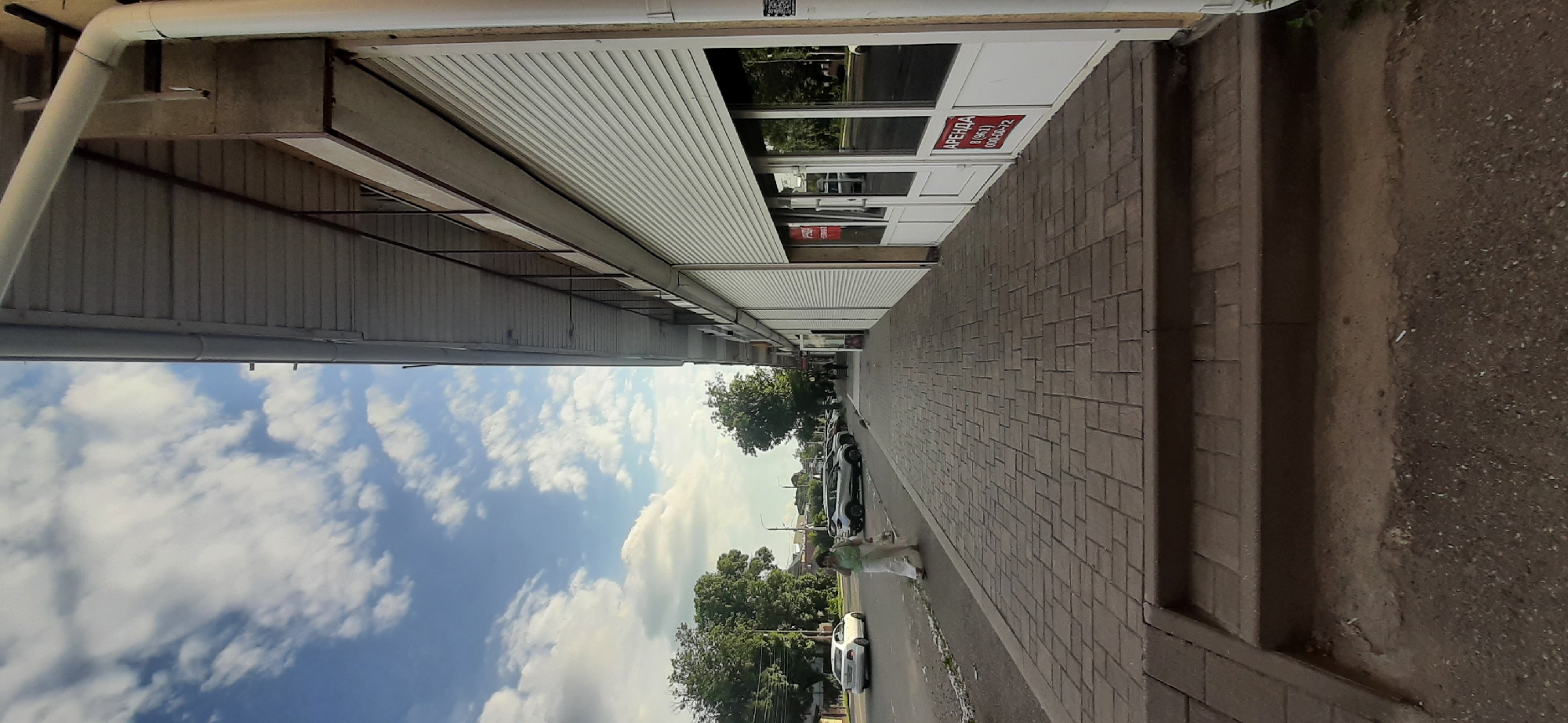 A low-angle, wide shot captures a street scene in Yessentuki, Russia, on a bright, cloudy day. The perspective is from a sidewalk, looking outwards towards a commercial building and a street lined with parked cars.
The commercial building features a facade with white metal roll-up doors and glass windows. A red sign with white lettering that reads "АРЕНДА" (ARENDA, meaning "RENT") is prominently displayed on one of the glass doors, along with a phone number. This suggests commercial activity or availability for lease. The building has a covered walkway or awning extending outwards.
The sidewalk is paved with textured brown bricks laid in a regular pattern. Adjacent to the sidewalk, there's a strip of asphalt or road surface where several cars are parked. Further down the street, trees with lush green foliage line the road, indicating a pleasant weather condition.
A person, appearing to be a woman dressed in light-colored clothing, walks on the sidewalk. The sky above is a vibrant blue with scattered white clouds, typical of a clear, sunny day. The overall impression is a typical urban street scene, likely during daylight hours, with a focus on commercial spaces and everyday street life. The image's composition emphasizes the vastness of the sky and the linear perspective of the street and building.