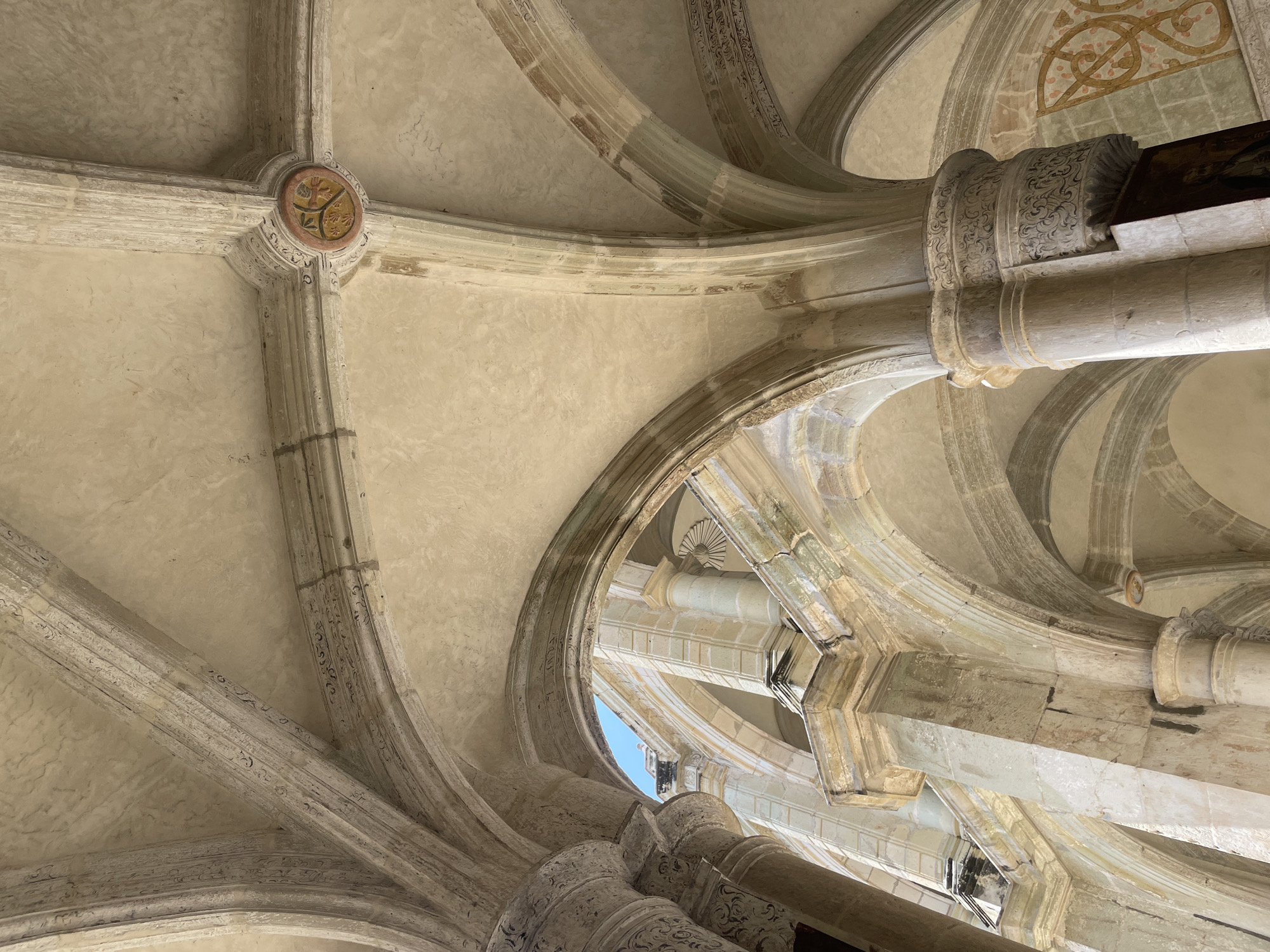 The image shows a view of the ceiling of the Templo de Santo Domingo in Oaxaca City, Mexico. The ceiling is an intricate network of stone arches and columns, with ornate carvings and embellishments. A small patch of blue sky is visible through an opening in the ceiling. The stonework has a weathered appearance, reflecting the building's age. There are some faded paintings at some point along the ceiling.