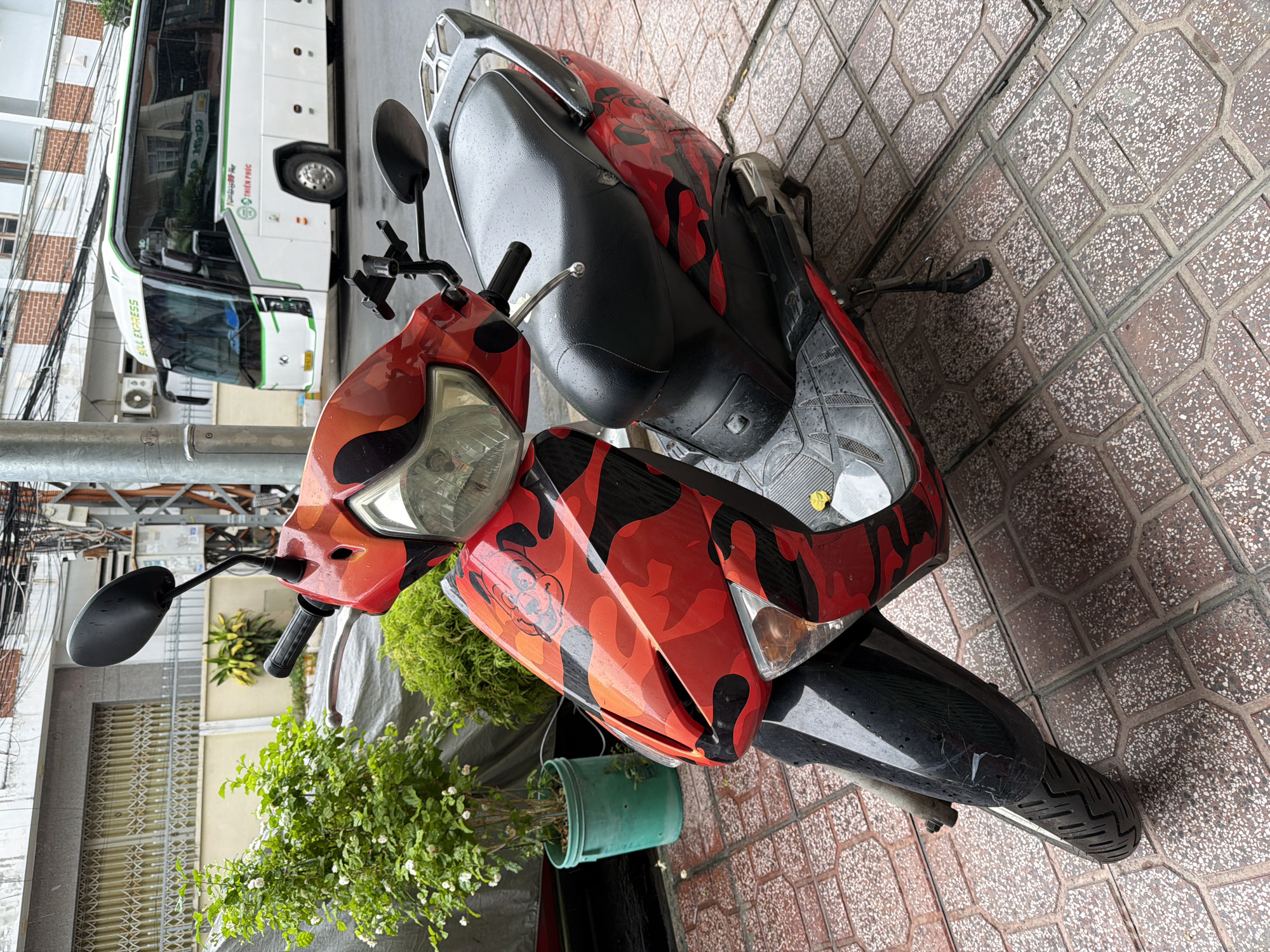 A red and black camouflage patterned scooter is parked on a paved sidewalk in Nha Trang, Vietnam. The scooter has a black seat and chrome accents. Behind the scooter, a white tour bus with green trim is visible on the street. The bus has "SOUL EXPRESS" and other text in Vietnamese written on its side. The sidewalk is made of red and gray tiles. A small green potted plant with white flowers is to the left of the scooter. A building with a grey facade and a metal gate is in the background. The scene appears to be an outdoor urban environment.
