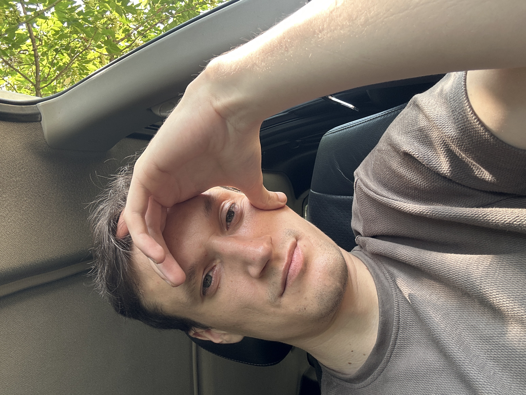 A selfie is taken from the perspective of a person sitting inside a vehicle, possibly a car, based on the visible interior. The person, appearing to be a young man with short, dark hair and a slight stubble, is looking directly at the camera with a pensive expression. His right hand is raised to his forehead, with his fingers resting on his brow. He is wearing a light brown, textured crewneck shirt. Sunlight streams in from the right side of the frame, illuminating his face and arm, and casting shadows. Outside the car window, lush green foliage suggests an outdoor setting, possibly in a rural or suburban area with trees. There is no specific indication of the city of Makiv, Ukraine in the image.