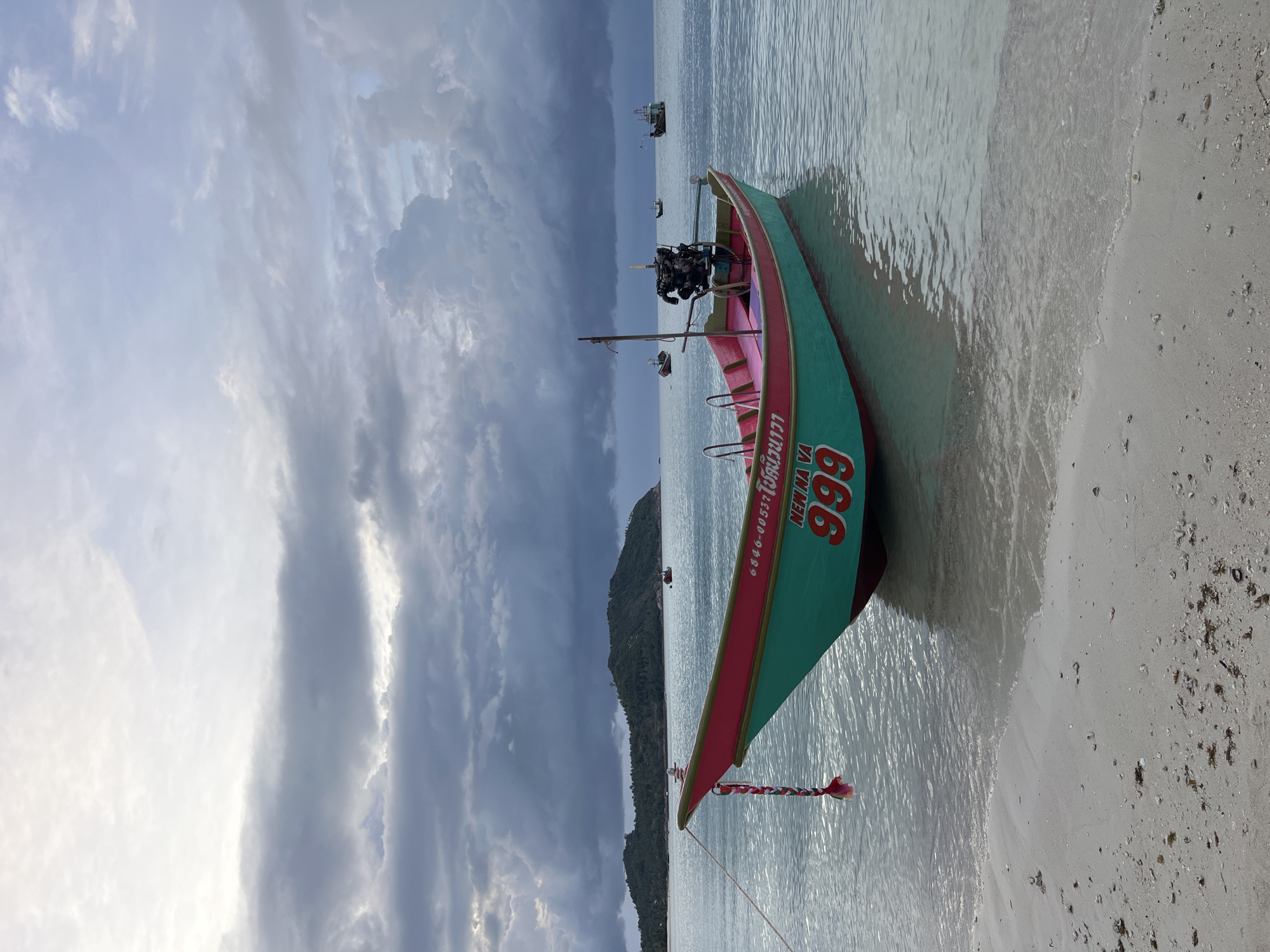 An eye-level, medium shot captures a Thai longtail boat docked near the shore of a beach on Ko Pha-ngan District, Thailand, under a dramatic, cloudy sky. The boat, painted teal with a pink interior, is tethered to the shore by a rope and has the number "999" painted in red on its side, along with some Thai script. A small outboard motor is mounted on the stern. The water is calm and clear, with gentle waves lapping at the sandy beach, which is littered with small shells and debris. In the background, a lush green island is visible in the distance, as are other boats on the horizon. The overcast sky suggests a moody atmosphere, possibly indicating an approaching storm. There are no people visible in the frame.