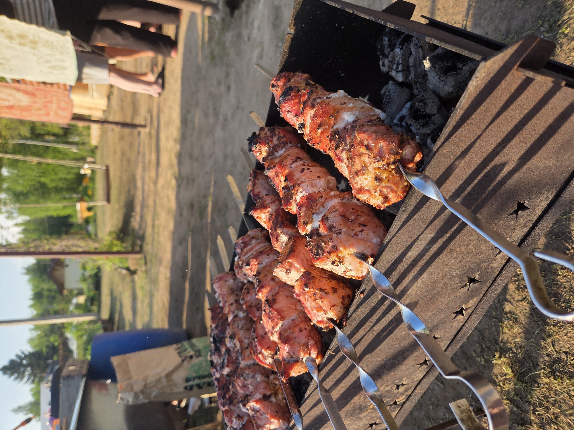 The image shows skewers of grilled meat cooking on a barbecue grill. The skewers are metal and have pieces of cooked meat on them. The grill is black and has charcoal inside. The background is blurred, but it appears to be an outdoor setting with trees and grass. Based on the user prompt, it is likely that the location is Krasnokamsk, Russia. Some other objects in the background include a barrel. Some people can be seen behind the grill.