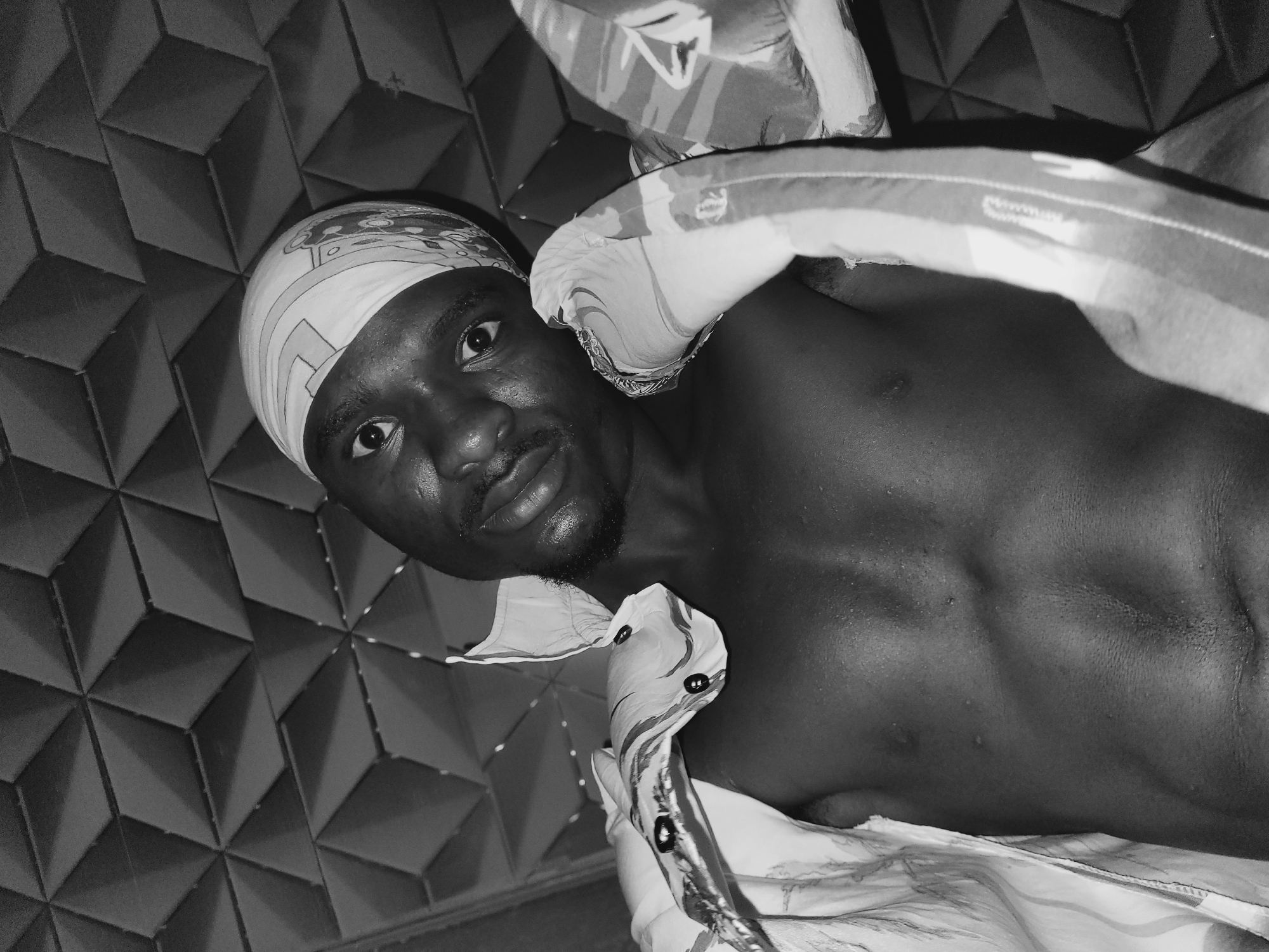 In a close-up, black and white, wide-angle shot, a person lies on their back in what appears to be a bedroom. The background features a textured wall with a repeating geometric pattern, possibly wallpaper or a decorative panel. The person, who appears to be male, is looking directly at the camera. They are wearing a patterned headscarf and their upper torso is visible. The body is glistening, suggesting sweat, and is partially covered by patterned fabric, possibly a duvet or bedsheets. The lighting appears to be overhead, casting shadows and highlighting the contours of the person's face and chest. The overall impression is intimate and possibly candid.