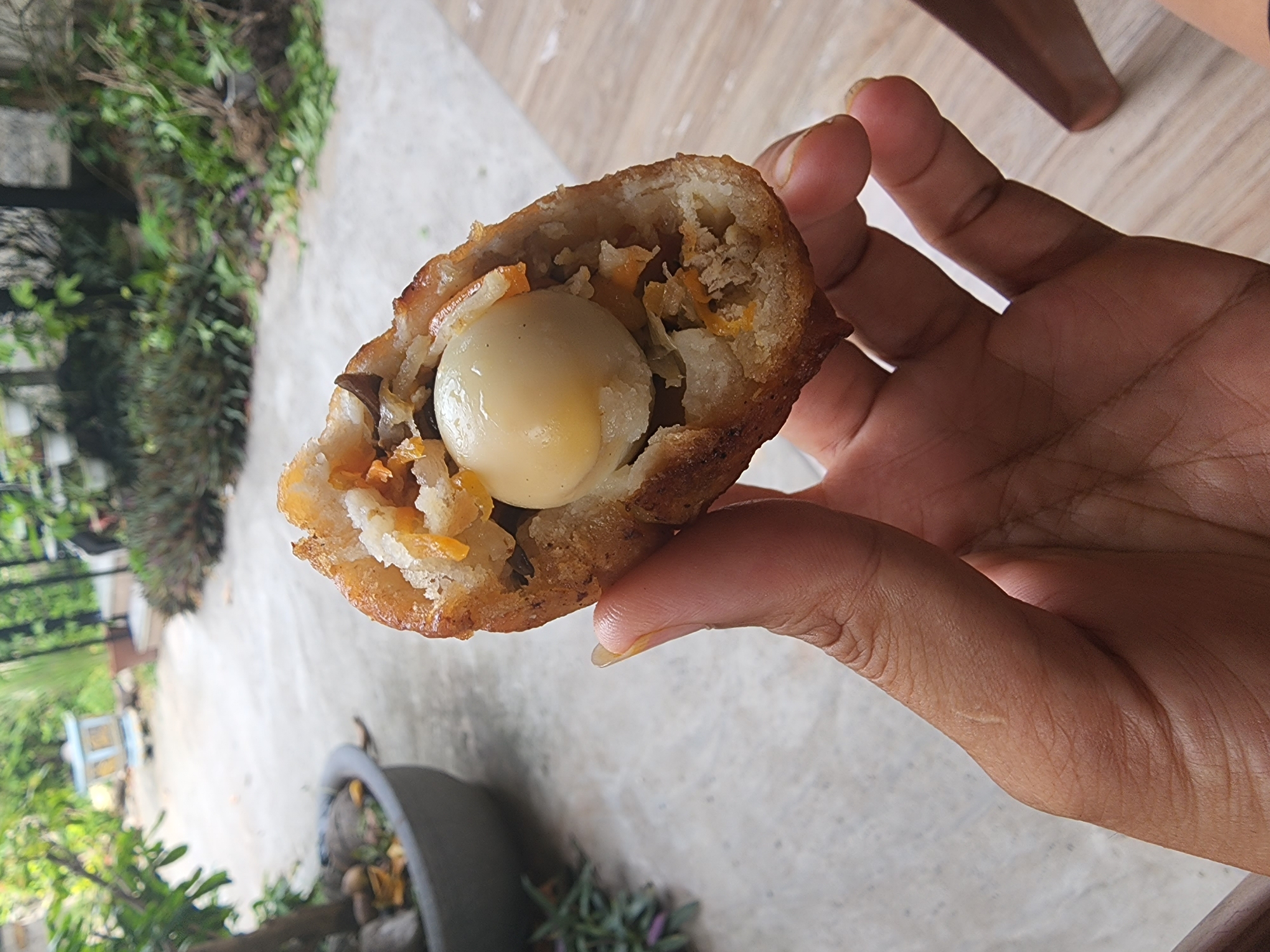 A close-up shot captures a hand holding a piece of fried food that appears to be a spring roll or similar snack, split open to reveal its filling. The filling contains what looks like shredded vegetables, possibly carrots or yams, minced meat, and a whole, small boiled egg, likely a quail egg, at the center. The exterior is golden brown and crispy from frying. The background is blurred, showing an outdoor setting with concrete paving, some greenery, potted plants, and a light-colored wooden surface. The scene is outdoors, suggesting a casual dining or snack moment. The specific location within Phan Thiet Ward, Vietnam, is not explicitly identifiable from the visual elements alone, but the outdoor setting and the food type are consistent with casual eateries or home environments in Vietnam.