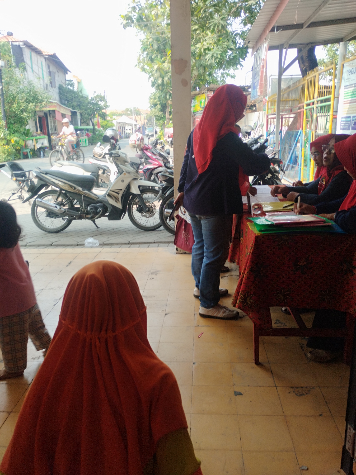 This is a medium shot taken from a low angle, showing a street scene in Surabaya, Indonesia. In the foreground, two children wearing orange headscarves are looking away from the camera. Behind them, a woman in a dark blue top and jeans, also wearing an orange headscarf, is standing next to a table covered with a red, patterned cloth. On the table are several folders and papers, and the woman appears to be interacting with individuals seated behind the table. To the left of the woman, several motorcycles are parked on a tiled walkway. Further in the background, a street is visible with buildings, trees, and a person riding a bicycle. The overall scene suggests an outdoor event or gathering.