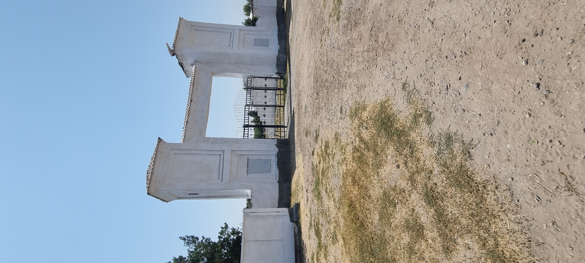 The image depicts a white arched gateway with two flanking square towers, a tiled roof, and black wrought iron gates in front of it. The ground in front of the gateway is a mixture of dry grass and gravel. A white wall extends to the left of the gateway. The sky is clear and blue, suggesting a sunny day. The overall scene appears to be an entrance to a property or facility in a dry, possibly rural, environment.