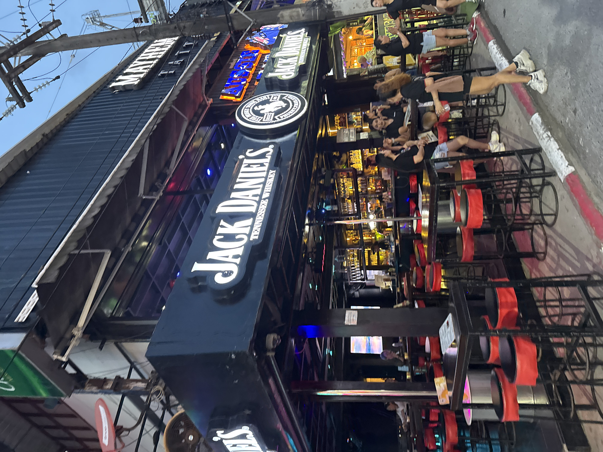 In Patong, Thailand, a street-level view captures a lively scene outside a bar. The bar, prominently featuring "Jack Daniel's Tennessee Whiskey" signage, has a dark exterior with glowing neon lights illuminating the interior where bottles are displayed on shelves behind the counter. Red bar stools and high-top tables are arranged outside, some occupied by patrons. The street itself is paved with a red and white striped curb. Several people, mostly women, are visible, some standing and conversing, others seated at tables or walking past. A "Mayhem Tattoo" sign is also visible on the upper facade of the building. The time of day appears to be dusk or early evening, indicated by the dimming natural light and the illuminated signs.