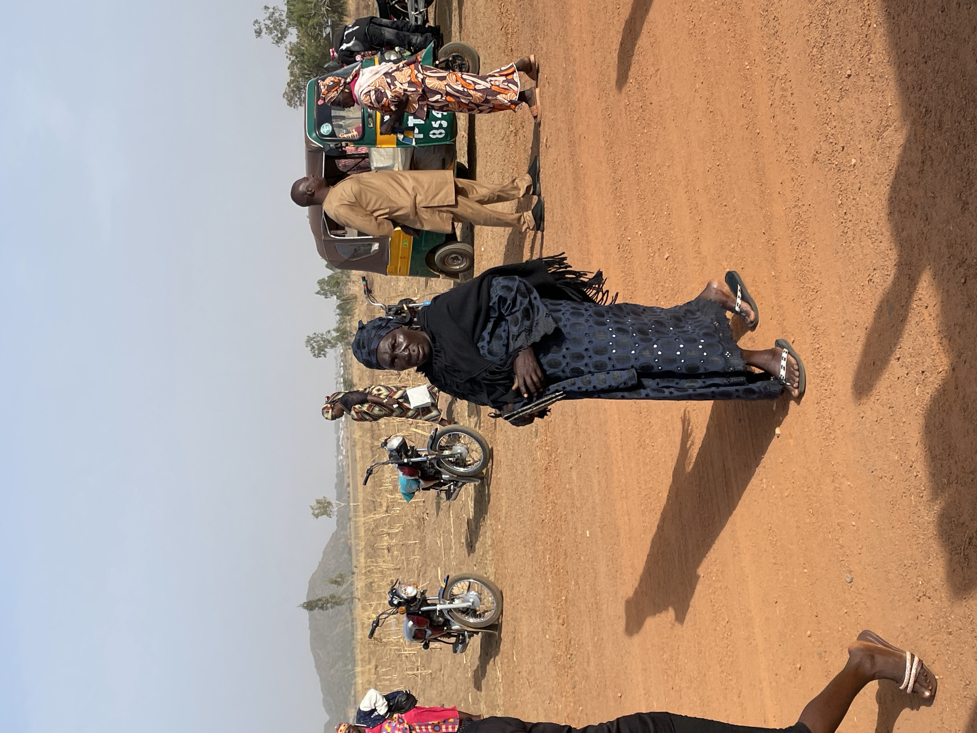 The image appears to be taken in a rural area in Nigeria, likely near the city of Du. The environment is dry, with reddish-brown dirt, sparse vegetation, and a distant hill. Several people are present, including an elderly woman in a dark blue patterned dress and a black head covering, standing in the foreground. She holds what appears to be several slender, dark objects in her hand. Behind her are two motorcycles parked on the dirt. Further back, a three-wheeled vehicle (likely a "keke" or auto-rickshaw), and two other individuals, including a man in a tan suit. The sky is clear and pale blue. There are other individuals in the background in the distance, and to the lower part of the frame are parts of other individual's bodies.