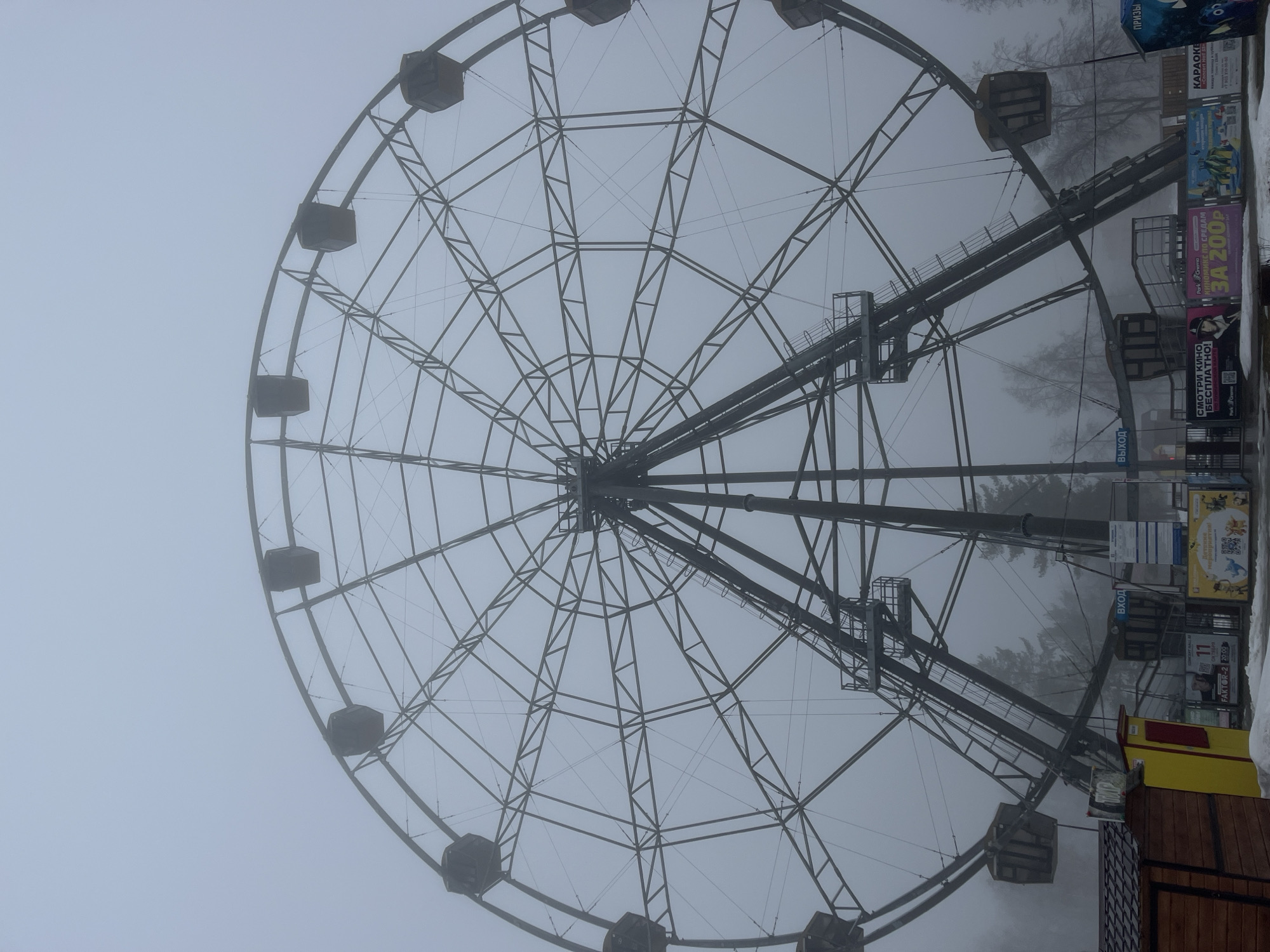 The image shows a Ferris wheel in a heavy fog or mist. The Ferris wheel's structure is visible, with the passenger cars spaced around the perimeter. The surrounding environment is obscured by the fog, but some advertisements and structures near the base of the Ferris wheel can be discerned. Text on the signs is partially legible, with references to "KARAOKE" and "ParkCinema." Based on the signs and other context clues, the location is likely Neftekamsk, Russia.
