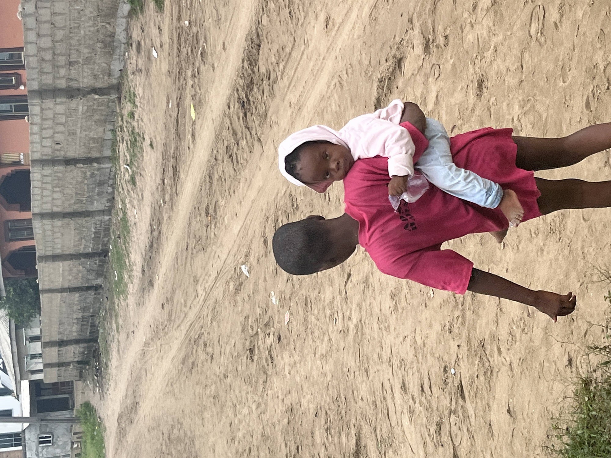 A child is being carried piggyback by another child on a dirt road in Badagry, Nigeria. The child on the back is wearing a pink hooded shirt and light blue pants and is looking directly at the camera. The carrier is wearing a pink shirt. The road is sandy and unpaved. In the background are buildings, some appearing under construction. There is some sparse greenery around the buildings.