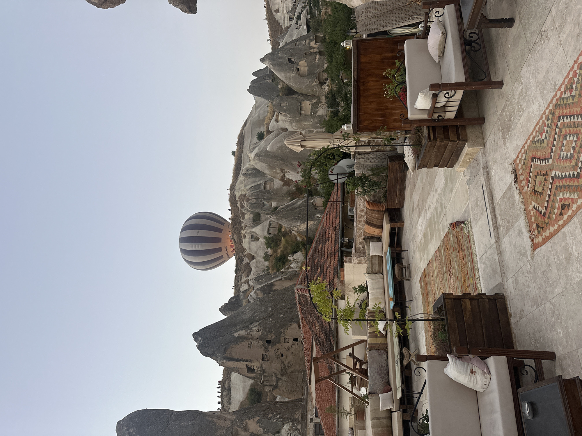 The image depicts a rooftop terrace in Göreme, Turkey, overlooking a landscape of unique rock formations. In the background, a hot air balloon with white and blue stripes is ascending into a clear, pale sky. The terrace is furnished with comfortable seating, including a long sofa with multiple cushions, a smaller bench with two cushions, and a single chair. Several woven rugs are placed on the paved floor. Wooden planters with small trees and plants add greenery to the space. A wooden swing set is visible on the left side of the terrace. The architecture of Göreme, characterized by its fairy chimneys and cave dwellings carved into the rock, is prominently featured in the midground and background. The overall scene suggests a serene morning atmosphere, likely during sunrise, given the presence of the hot air balloon.