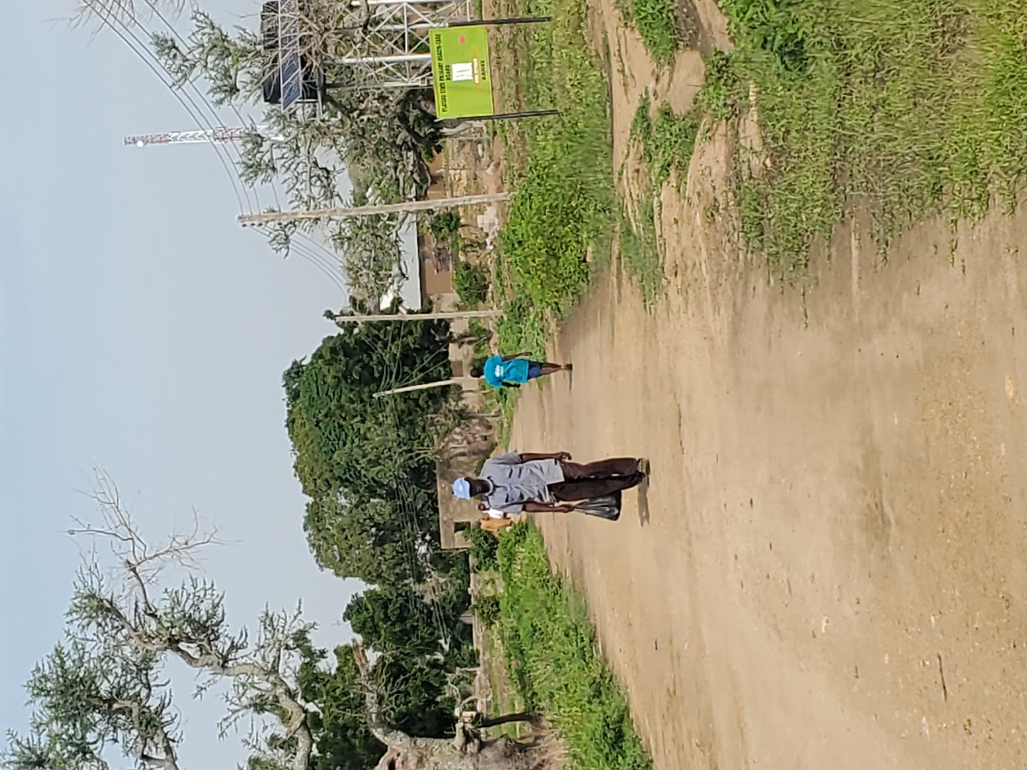 The media file depicts an outdoor scene with two individuals walking on a dirt road. The person in the foreground, closer to the viewer, is a man wearing a grey short-sleeved shirt, brown pants, and a blue cap. He is carrying a black bag in his right hand. Behind him and further down the road, a second person, appearing to be a child, is wearing a blue t-shirt and shorts. They are walking away from the camera.
The setting is rural, with vegetation and trees lining the sides of the road. In the background, a large metal tower with electrical lines is visible, suggesting the presence of infrastructure. A green sign with some text is also present near the tower. The road itself is dusty and appears to be unpaved. The sky is overcast, suggesting a potentially cloudy or hazy day.
The activity is simply walking along the road. There are no overt indications of a specific event or strong emotions. The time of day appears to be daytime, but the lighting is diffused due to the overcast sky. The location is not definitively known, but the presence of the tower and the general landscape suggest a more remote or developing area.
Visible text on the green sign includes: "PLACING POST PRIMARY HEALTH CARE" and "BOARD", and "KAHEL". The orientation of the image is rotated, with the road running vertically from bottom to top and the sky at the top.