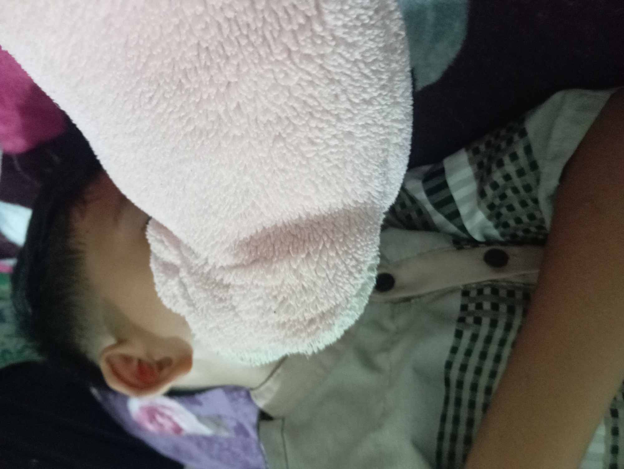 The image shows a close-up of a child sleeping, with a pink towel draped over their head and shoulder. The child appears to be lying on a bed with patterned bedding. The child is wearing a light-colored collared shirt with a checkered pattern. The setting appears to be indoors, likely a bedroom. There is no discernible information about the city of Rejosari, Indonesia in the image.