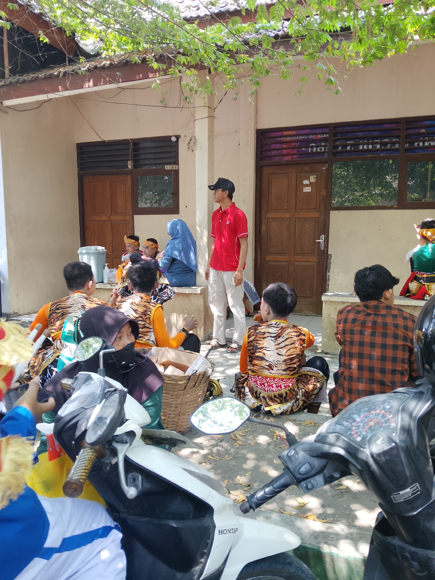 The image depicts an outdoor scene in Balun, Indonesia, likely a schoolyard or community gathering space. In the foreground, two motorcycles are parked. The one on the right is a black scooter with its handlebars and dashboard visible. The one on the left is a white scooter, primarily showing its front fender and a portion of its body.
Several individuals are present. A man in a red polo shirt and light-colored pants stands near a wooden double door, facing towards the right of the frame. He appears to be observing the group. To his left, a group of children, likely boys, are seated. They are all wearing similar costumes featuring tiger stripes on orange vests and patterned bottoms. Some are facing away from the camera, while others are looking around. One child is wearing a blue headscarf.
Further to the right, a person in a red and black plaid shirt is seated, facing away from the camera. In the background, the building has beige walls and brown wooden doors and windows with louvers. Green foliage from a tree hangs over the roof, casting dappled shadows on the ground. A woven basket is also visible among the seated children. The overall impression is of a gathering, possibly a cultural event or performance preparation, given the distinctive costumes.