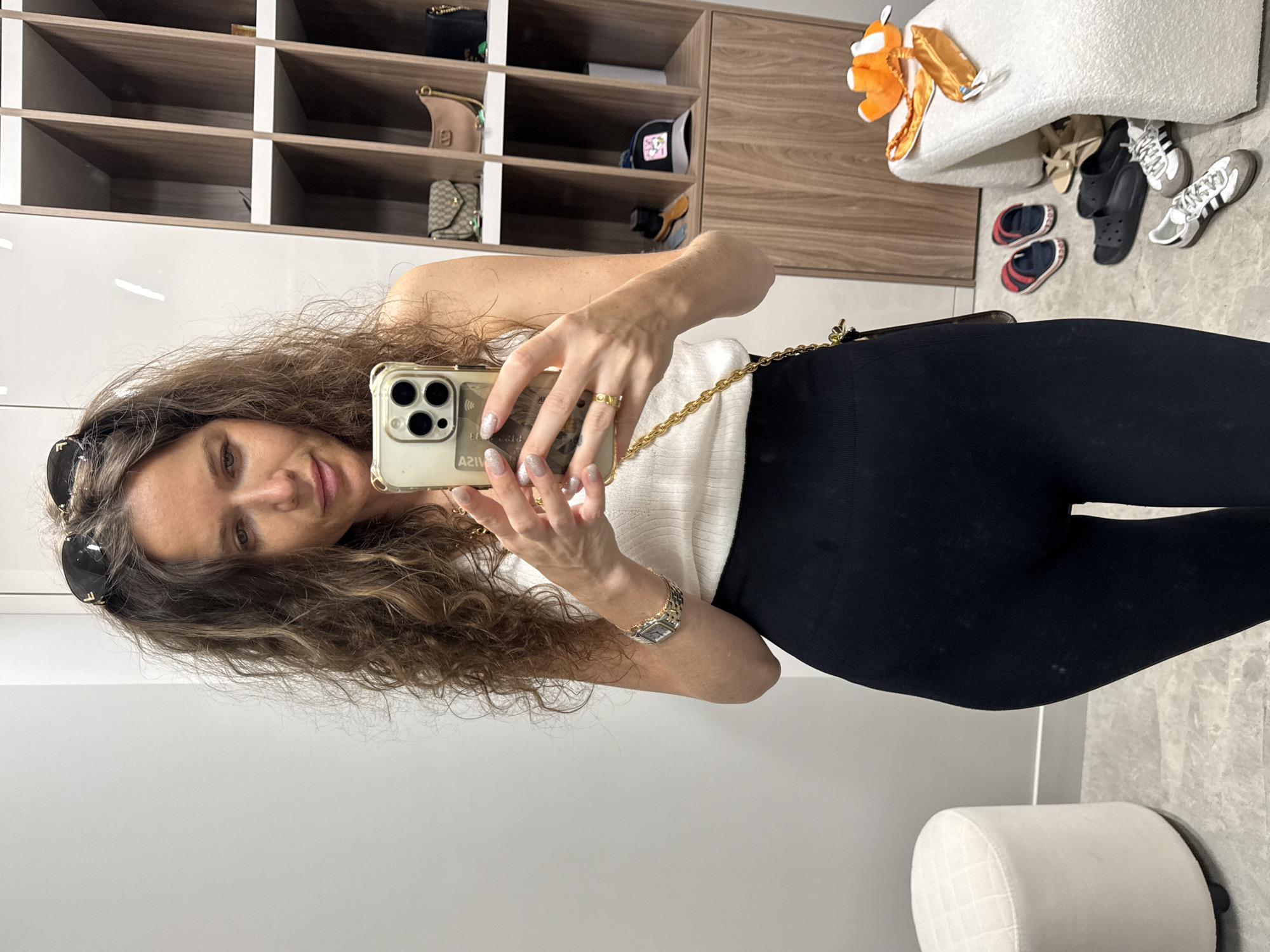 A woman stands in a room and takes a mirror selfie. She has long, curly brown hair, and is wearing a white tank top, black leggings, and sunglasses on her head. She is wearing a watch on her left wrist and a ring on her right index finger. Her phone is held in her right hand, and it is in a light-colored case with a gold chain strap. In the background, there is a white stool to the left and a shelving unit with bags and hats to the right. There are also several pairs of shoes on the floor near the stool and shelving unit.