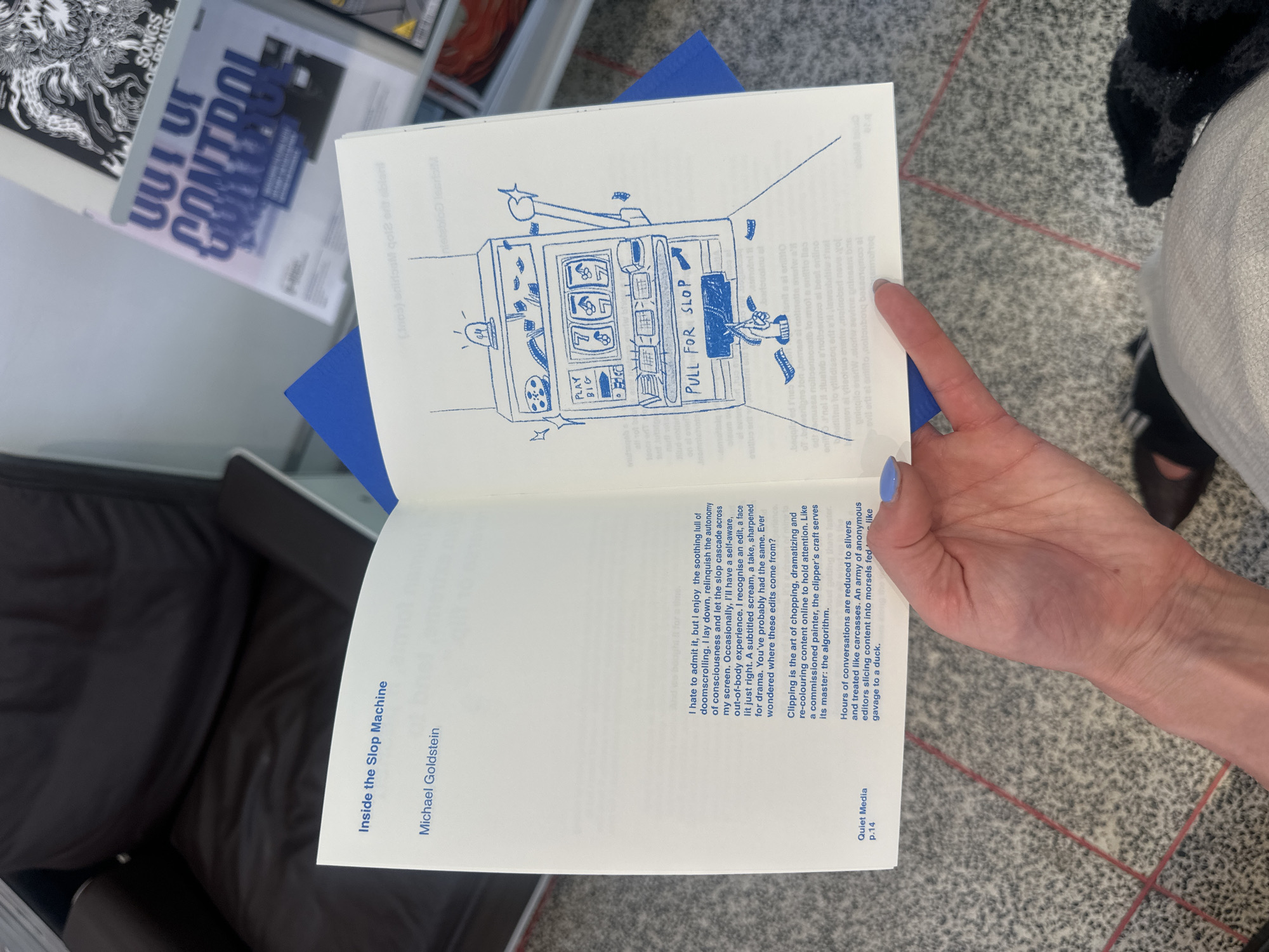 A person is holding open a book titled "Inside the Slop Machine" by Michael Goldstein. The book's pages are white with blue illustrations and text. The right page features a drawing of a slot machine, labeled "PULL FOR SLOP." The left page contains text describing doomscrolling, editing, and content creation. In the background, there are magazines on a shelf, a brown leather chair, and a patterned rug on the floor. The image was taken indoors, likely in a bookstore or home.