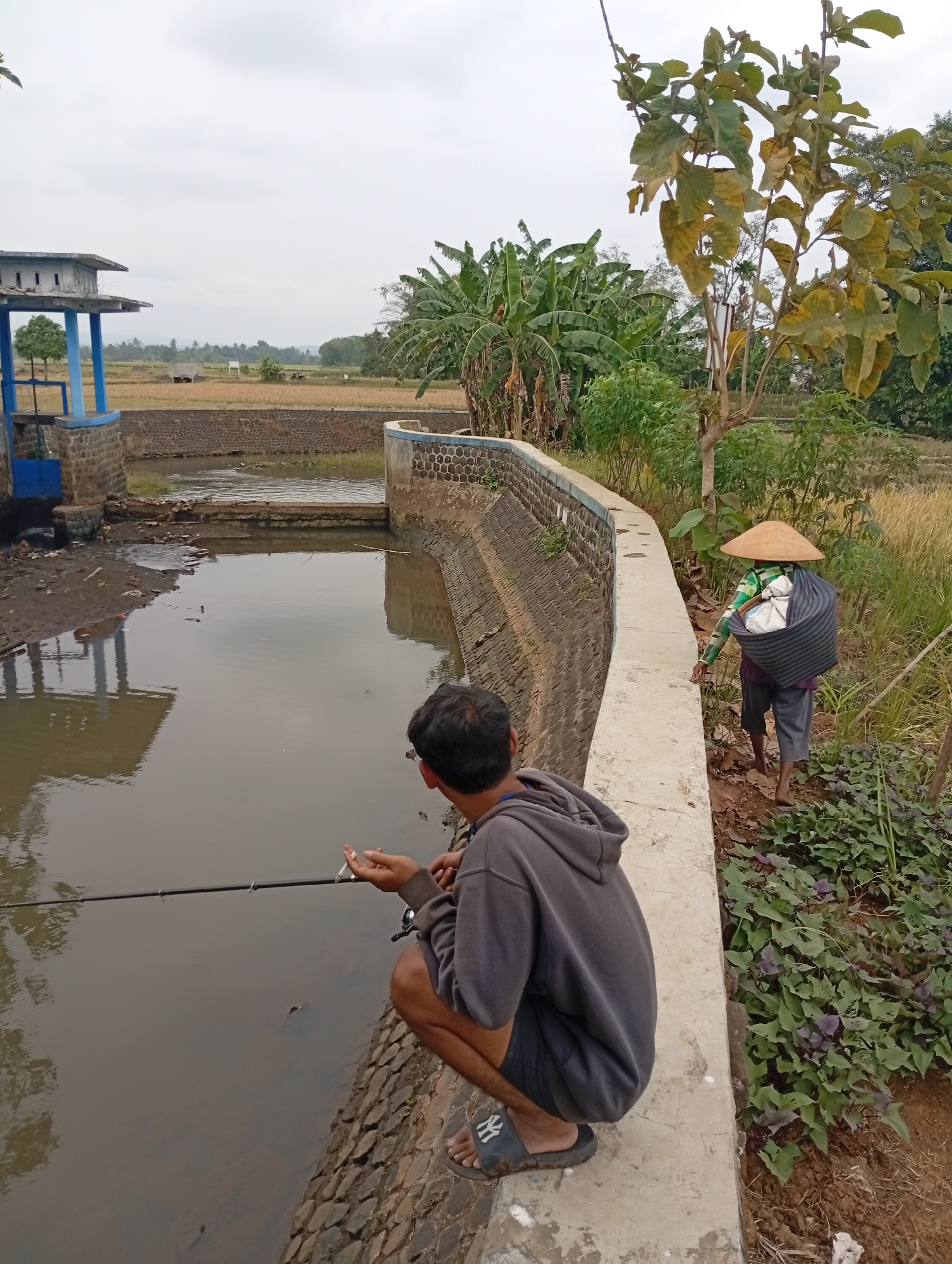 In an outdoor scene, two individuals are present near a body of water, possibly a canal or pond, with a rural landscape in the background. In the foreground, a person is crouched down, facing the water and holding a fishing rod, with a cigarette in their right hand. They are wearing a dark gray hoodie and dark shorts. Behind them, to the right, another person walks away from the camera, carrying a large basket on their back. This individual wears a conical straw hat, a green and white striped shirt, and dark shorts. The area around the water is lined with stone walls, and there is vegetation, including banana trees and other plants, surrounding the scene. A small structure with a blue accent, possibly a water gate or control building, is visible to the left. The sky is overcast, suggesting a cloudy day.