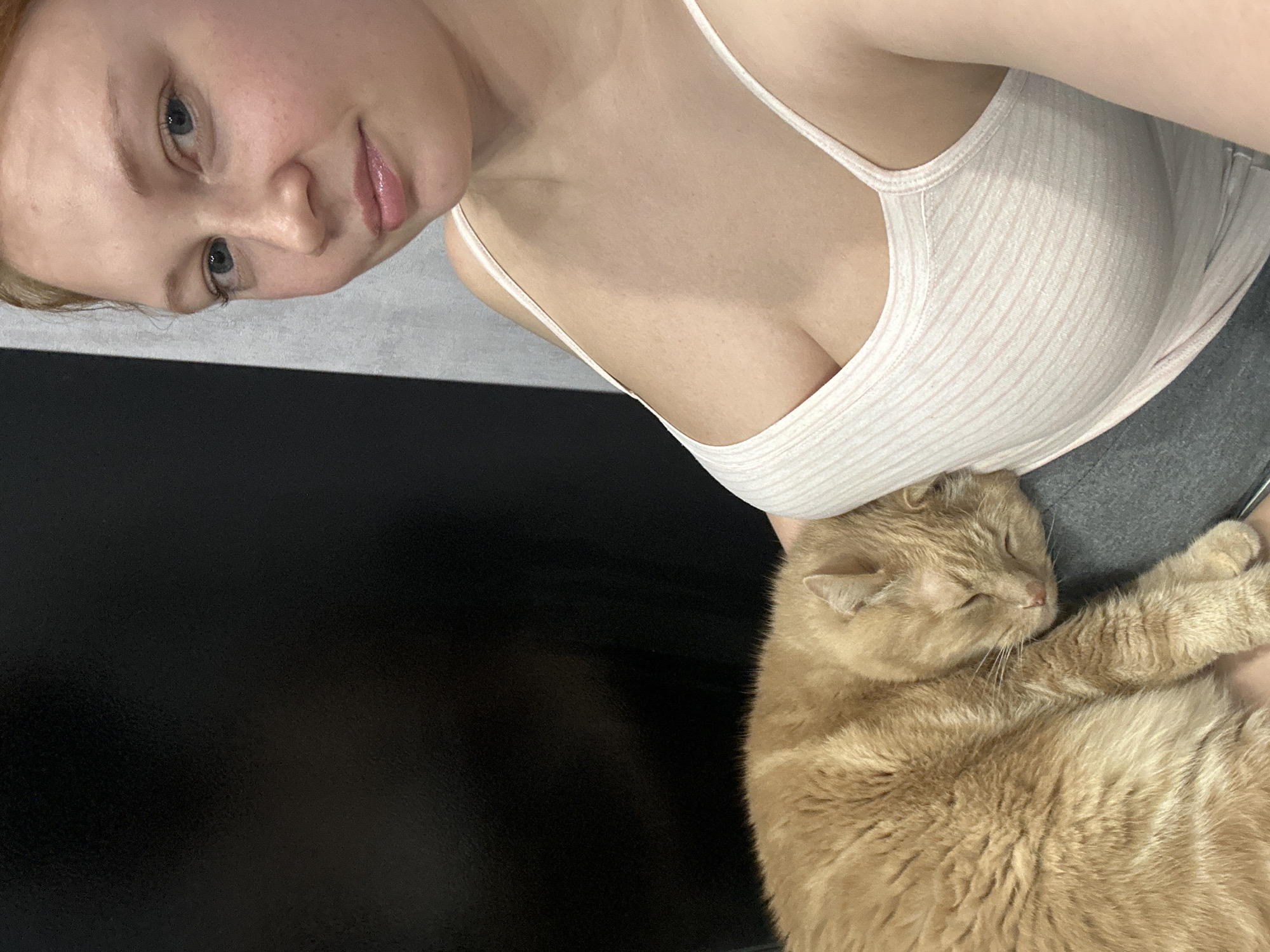 A fair-skinned woman with blonde hair and blue eyes is visible from the nose up, in the top left of the frame. She has pink lips. Her head is tilted slightly to the left. She is wearing a white tank top.
A large orange cat with closed eyes is curled up on her lap.
In the background, there is a black surface to the left, and a gray surface near the woman's lap.
There is no indication of the location being Monchegorsk, Russia.