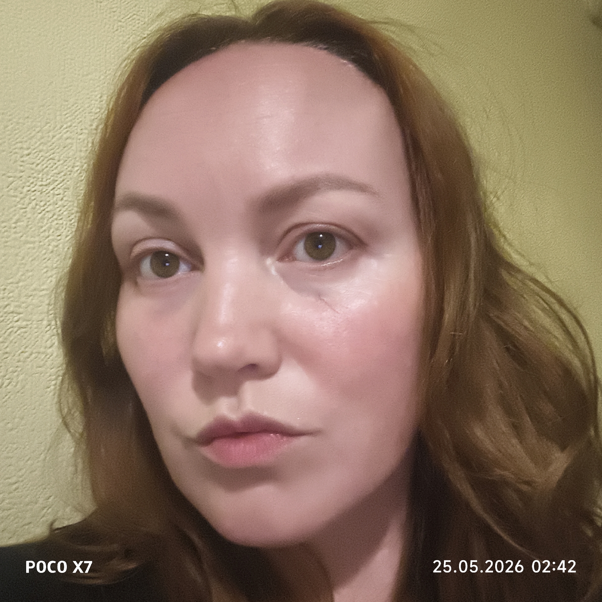 A close-up photograph depicts a woman with fair skin and auburn hair. She has light eyebrows and hazel eyes. A slight scar is visible near her right eye. The background appears to be a textured, light-yellow wall. The bottom of the photograph displays "POCO X7" and the timestamp "25.05.2026 02:42". The photograph appears to be taken indoors. Location in the city of Unknown, Unknown.