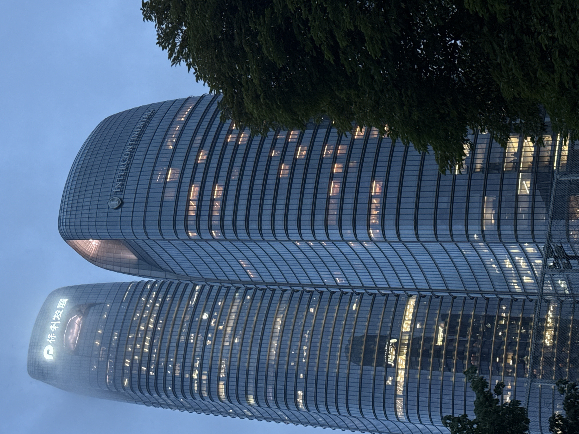The image shows two modern skyscrapers in Haizhu District, China, at dusk. The skyscraper on the left has a logo with Chinese characters, possibly "保利发展" (Poly Developments), illuminated at the top. The skyscraper on the right features the "INTERCONTINENTAL" logo. Both buildings are tall and have glass facades with many windows, some of which are lit from within, suggesting occupancy. The sky is a deep blue, indicative of twilight. A large, dark green tree partially obscures the right side of the right skyscraper. In the foreground, a chain-link fence is visible. The overall scene depicts a commercial urban environment.