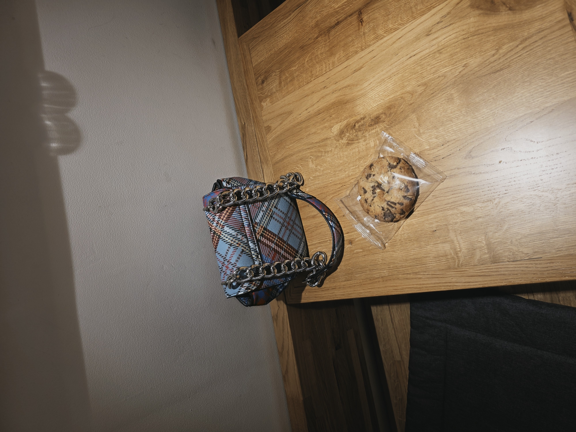 A plaid handbag with a silver chain strap sits on a light brown wooden table. Next to the handbag is a single chocolate chip cookie in a clear plastic wrapper. The table is placed against a light beige wall, which has a shadow cast upon it. The scene is illuminated by a light source positioned above and to the left, creating reflections on the wall. The environment appears to be indoors, possibly a cafe or a room in a residence, located in Saint Petersburg, Russia. The perspective is a high-angle shot, looking down at the objects on the table.
