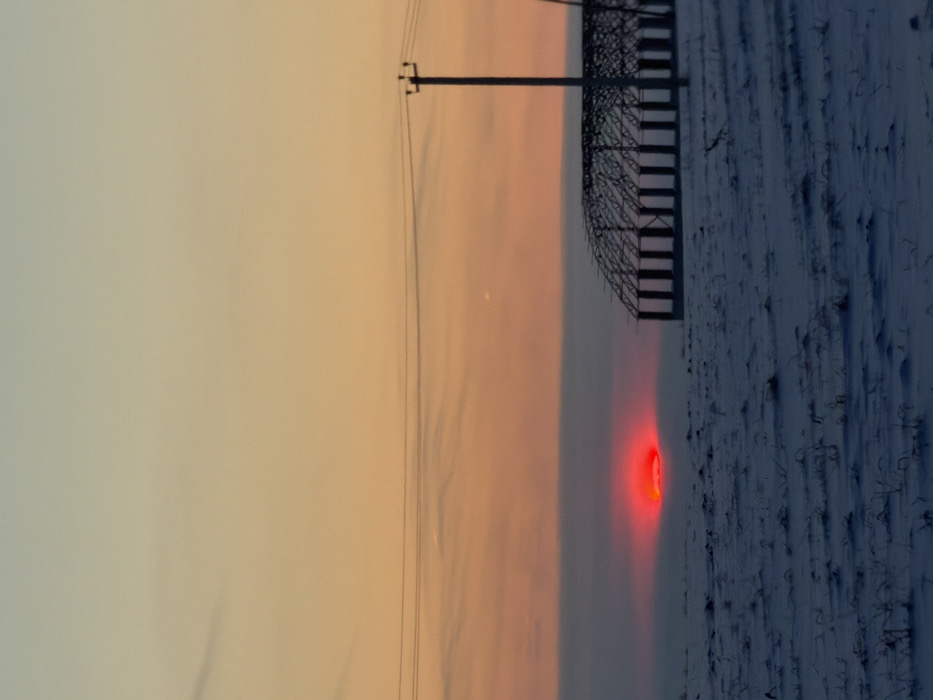 A landscape at dusk features a sky transitioning from light orange to muted gray. A telephone pole and the skeletal frame of a structure are visible against the fading light. A field covered in snow dominates the lower portion of the image, with sparse, dark vegetation poking through. A bright red, circular light source illuminates the snowy ground near the center, creating a striking contrast with the muted colors.