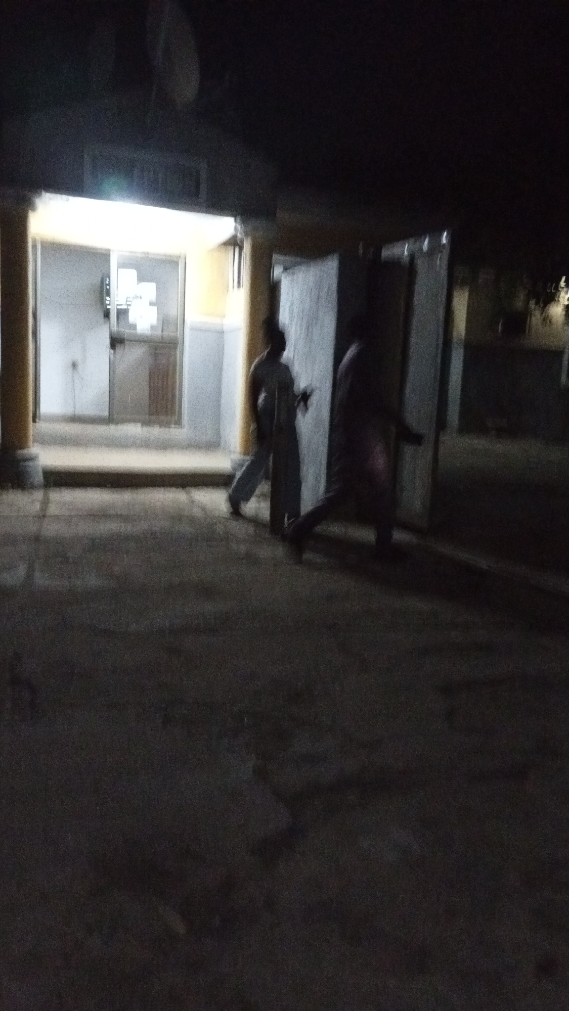 This image depicts a dimly lit outdoor scene at night in Adankolo, Nigeria. The primary focus is a brightly illuminated building entrance on the left, featuring a door with glass panels and a light-colored frame. Two sturdy pillars flank this entrance, which stands out against the dark surroundings. Above the entrance, a structure is visible, but any text on it is illegible due to overexposure and blur.
In the mid-ground, two human figures are present, both appearing to be in motion, indicated by slight motion blur. The figure on the left is wearing light-colored clothing and is seen walking. The second figure, positioned further to the right and partially obscured, also appears to be walking. Both individuals are located next to a series of large, light-colored rectangular objects, possibly partitions or panels, arranged vertically.
The ground in the foreground is paved, dark, and shows some texture or irregularities. The upper portion of the image and the background are consumed by darkness, though a faint outline of what resembles a satellite dish can be seen in the upper left. The overall scene is characterized by low light conditions, which contribute to graininess and reduced clarity, creating a somewhat mysterious ambiance. No readable text is visible in the image.