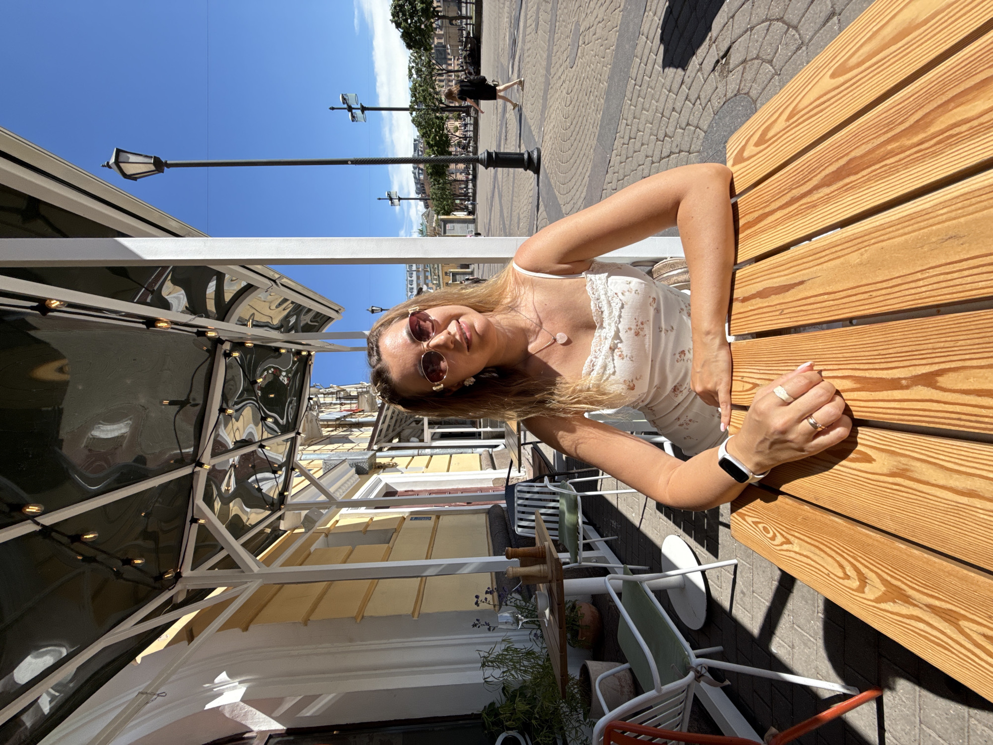 An outdoor, eye-level shot captures a young woman seated at a light-colored wooden slatted table at an urban outdoor cafe during daylight hours. She has long, light brown hair, wears dark sunglasses, a white floral patterned sundress with thin straps, a heart-shaped pendant necklace, rings on her fingers, and a white smartwatch on her left wrist. Her arms are resting on the table as she gazes towards the viewer. Above her, a dark, translucent awning supported by a white metal frame is strung with exposed-bulb string lights, creating reflections of surrounding buildings.
To her left, another smaller wooden table is visible with a wine glass, a white cup, and two wooden spice grinders. White metal chairs with light green cushions are arranged around the tables, alongside potted green plants and one with small purple flowers. The cafe is situated alongside a building with a yellow facade and white architectural trim.
Beyond the cafe patio, a paved pedestrian square or street extends under a clear blue sky. A tall, black traditional-style streetlight is prominent in the mid-ground on the right. Further in the background, another person wearing a black dress walks across the paved area, and several more modern streetlights and trees are visible, along with buildings lining the distant edge of the square. Strong shadows indicate bright sunlight.