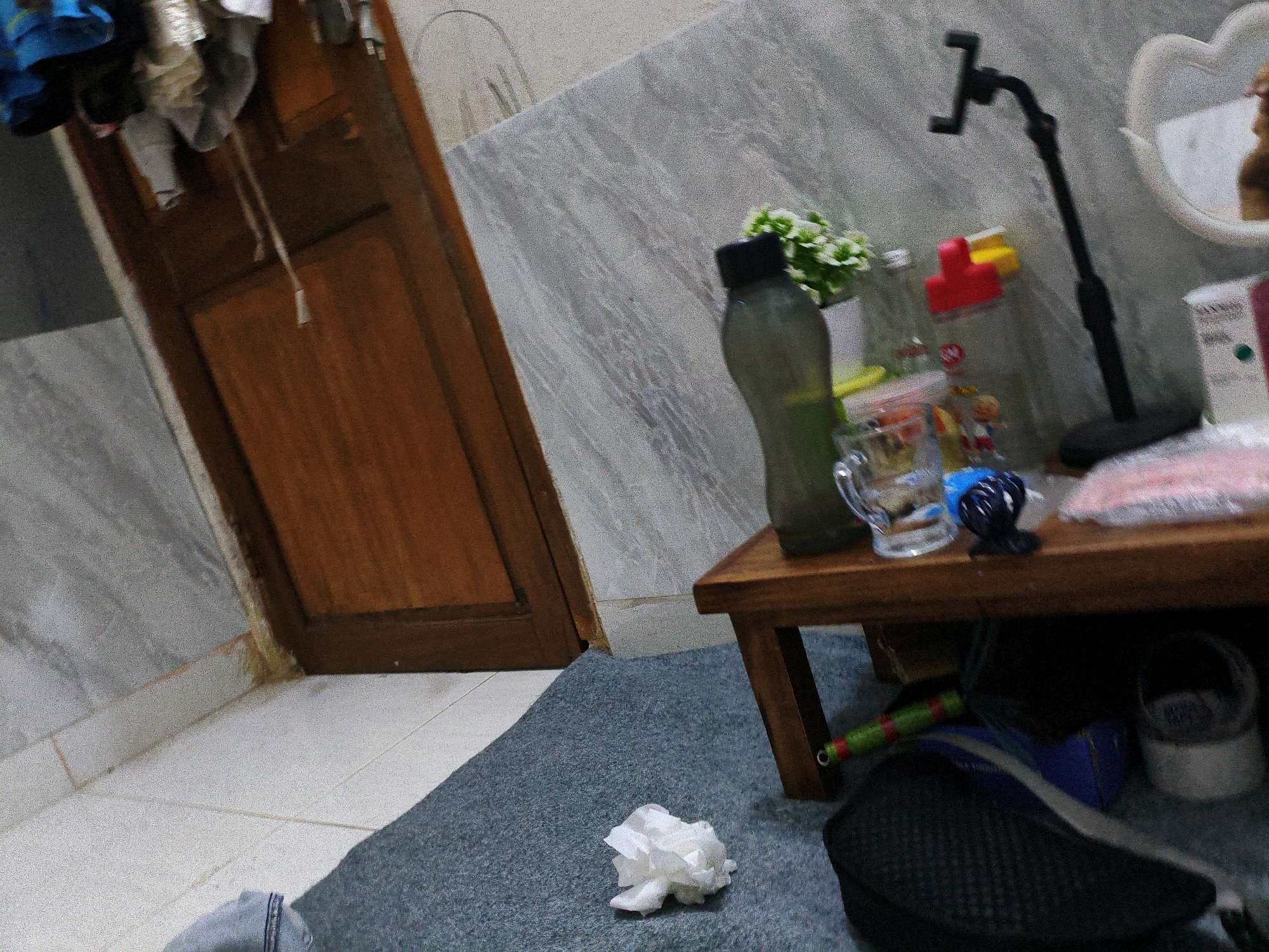 This image depicts an indoor scene, possibly a room in a house or accommodation in Karangtalun, Indonesia. In the background, a wooden door is visible on the left. The walls appear to be tiled with a gray, marble-like pattern. On a wooden shelf or table in the mid-ground, several objects are present: a dark-colored bottle, a clear glass with a handle, some colorful items including a red and yellow object, and a small plant with green leaves. A black stand with a phone holder is also visible on the shelf. In the foreground, a gray rug covers the floor, and a crumpled white tissue is on the rug. Below the shelf, a dark object, possibly a shoe or bag, is visible, along with a green and red striped object, likely a roller or stick. The overall impression is of a cluttered but functional living space.