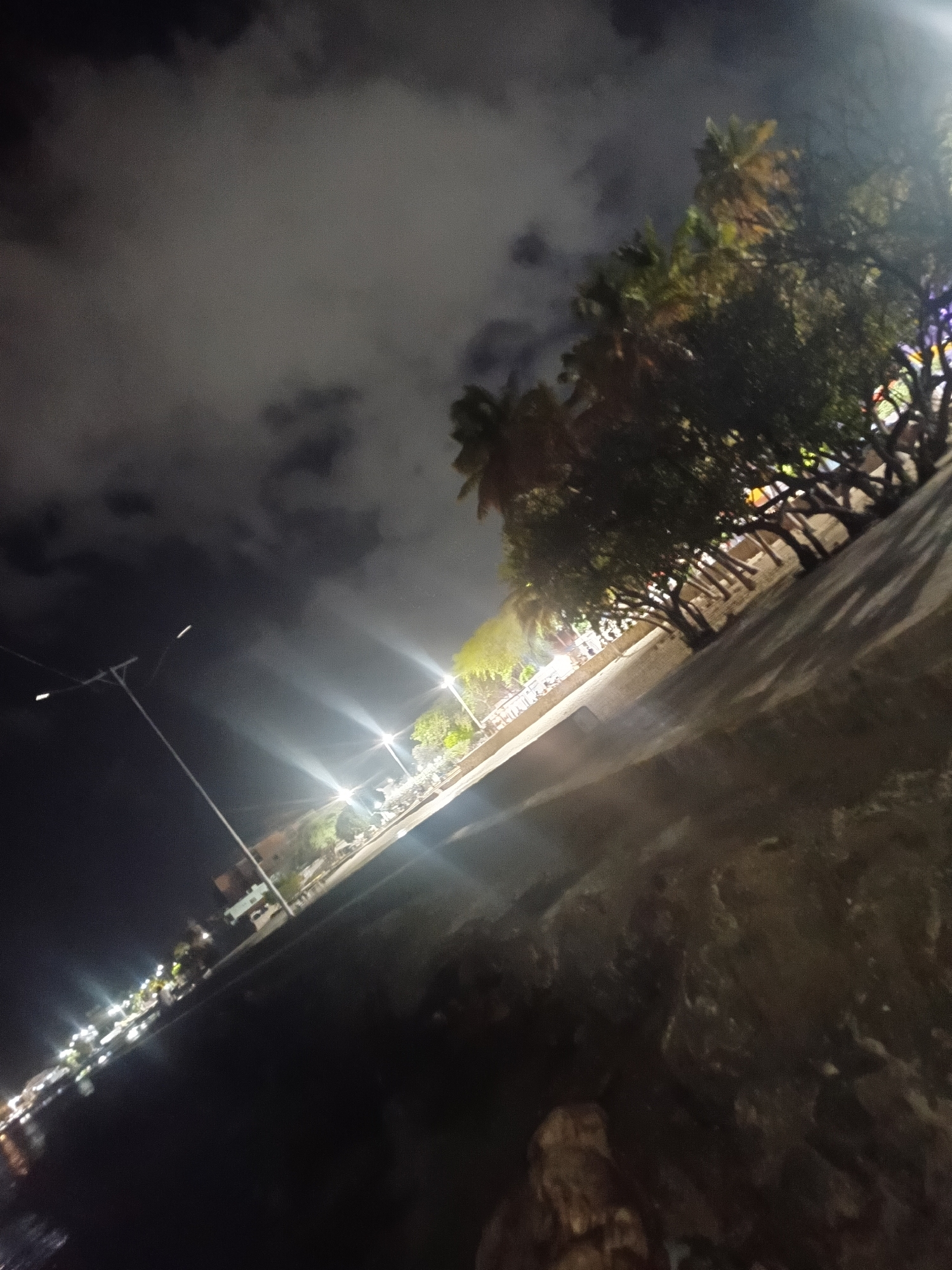 The image depicts a night scene in Juan Griego, Venezuela, likely along its waterfront or a well-lit promenade. The sky is dark with cloudy formations, and several streetlights are illuminated, casting bright beams onto the ground and illuminating the surrounding trees. The trees appear to be tropical, possibly palm trees, lining a pathway or road. In the distance, there are visible structures and more lights, suggesting a populated area. The foreground is dark, with what appears to be a textured surface, possibly a wall or embankment, at an angle. The overall impression is one of a lively urban night, illuminated by artificial lights against a dark sky.